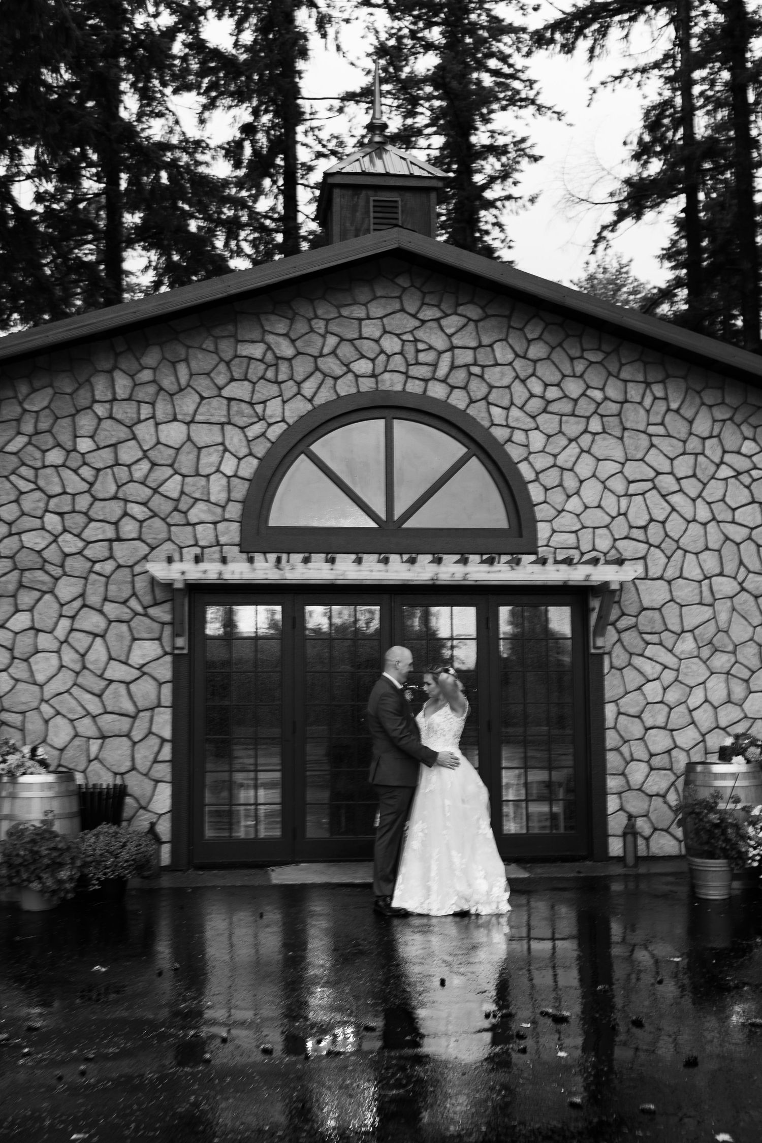 Bride and groom embracing in the rain outside a stone chapel venue in black and white.