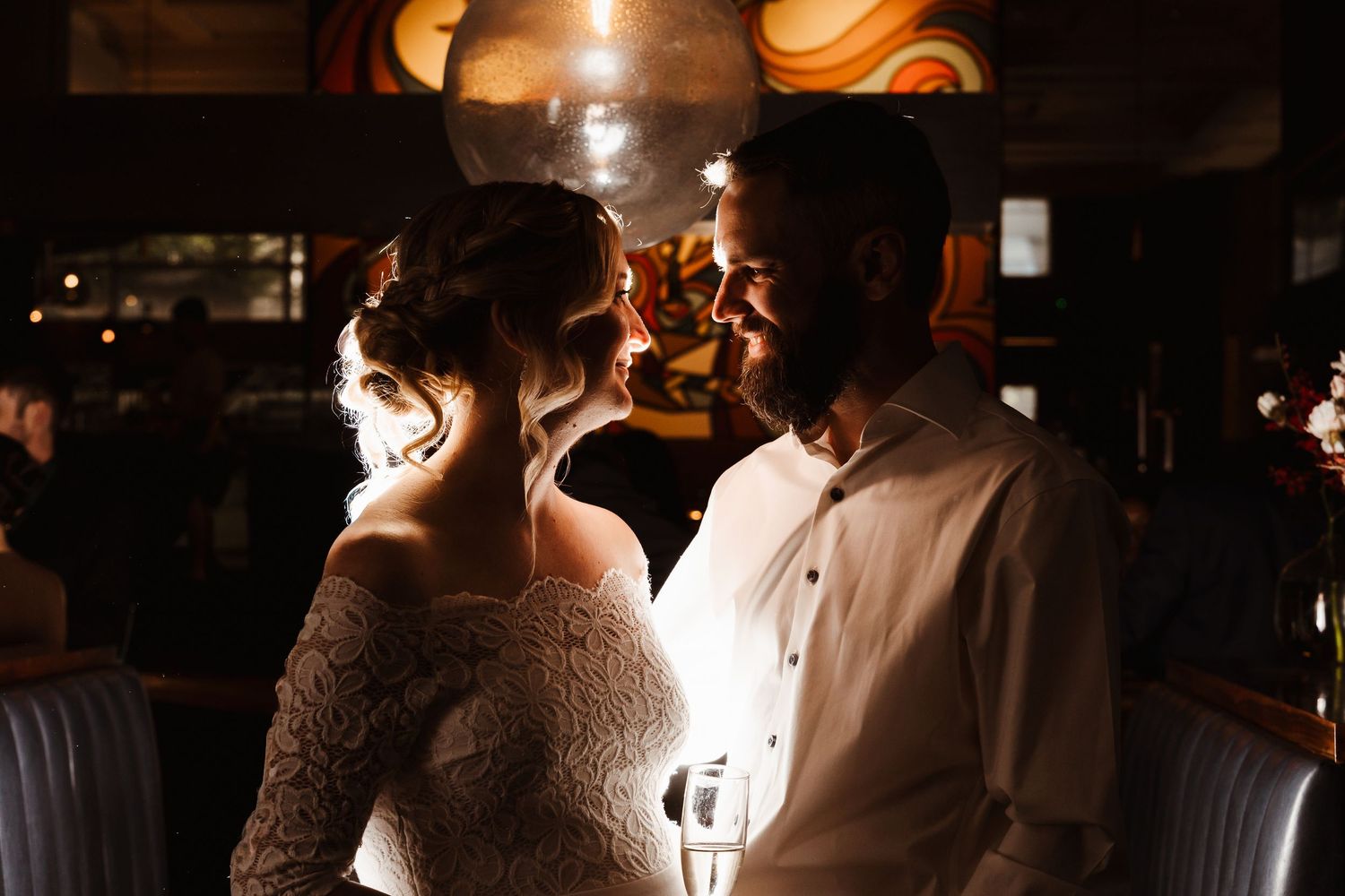 Romantic couple sharing an intimate moment at a dimly lit bar, bride in lace gown and groom in white shirt.