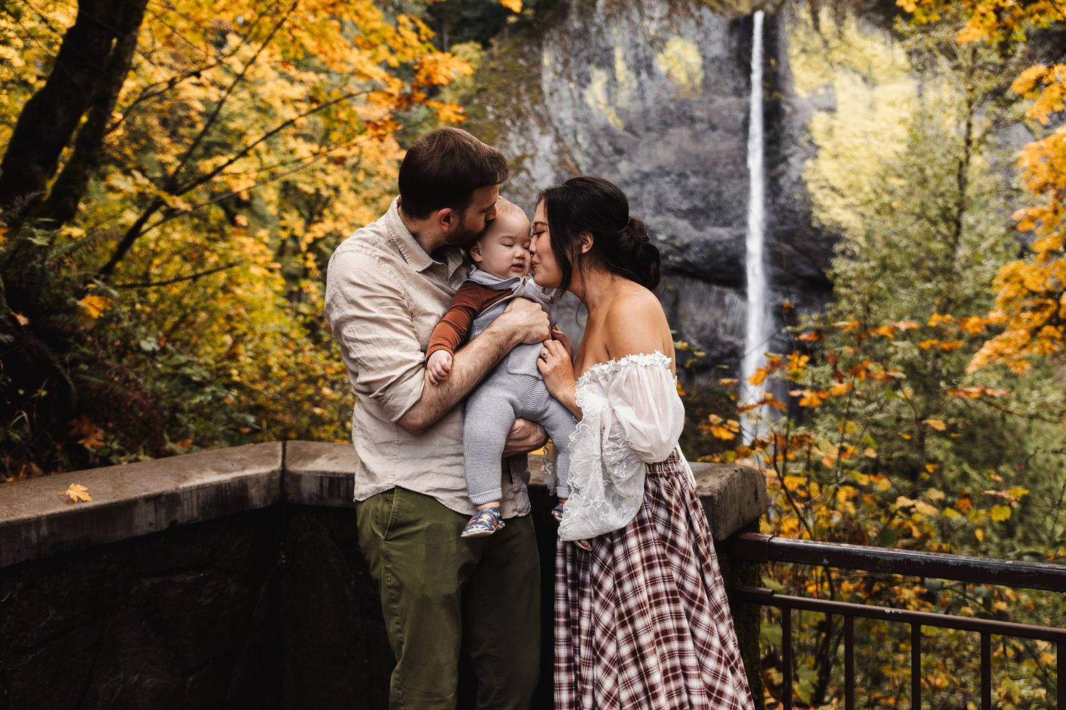 Family of three sharing a tender moment by a waterfall surrounded by vibrant autumn foliage.