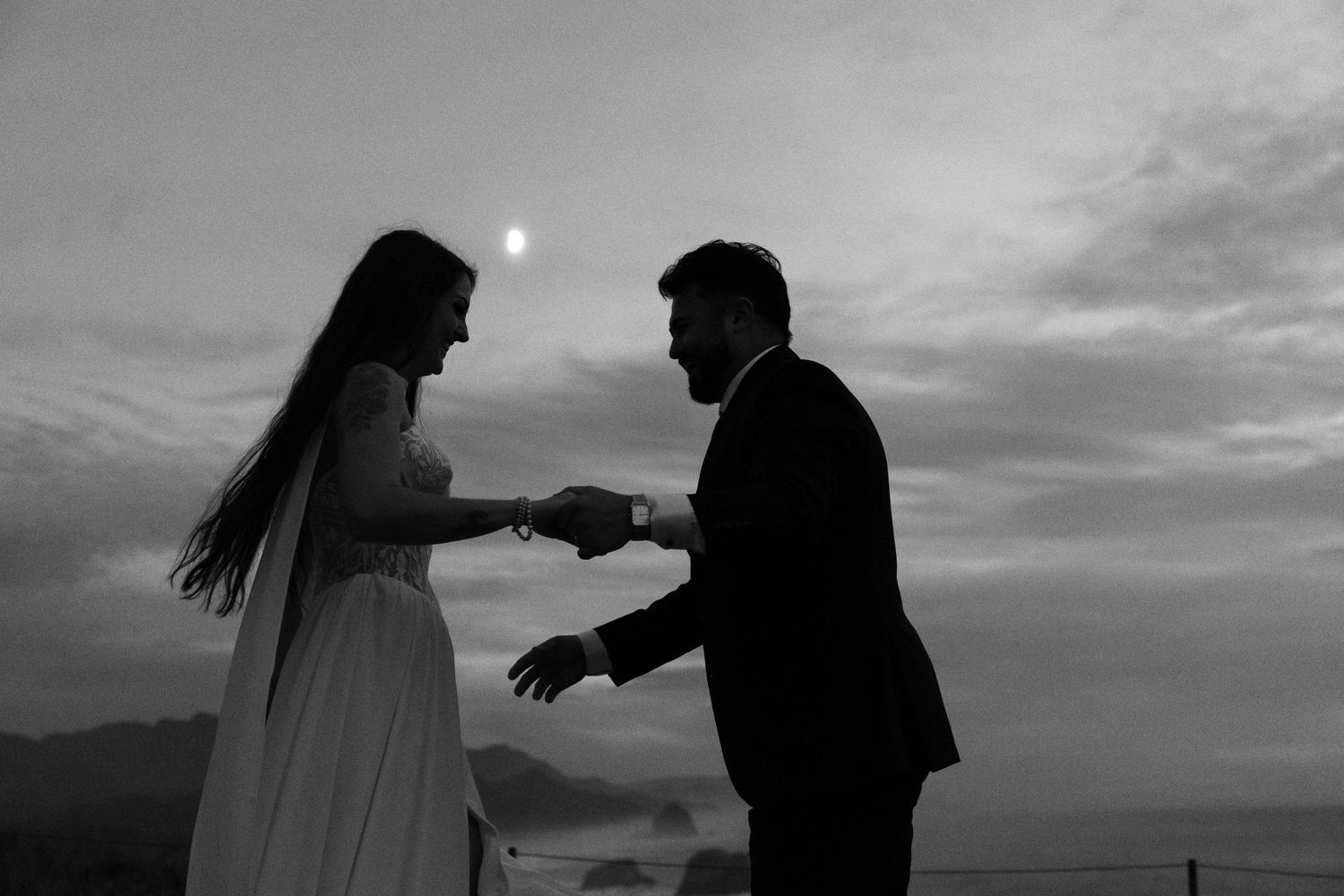 Silhouetted bride in a long veil and groom in a suit hold hands under a crescent moon at dusk in a black and white wedding portrait.