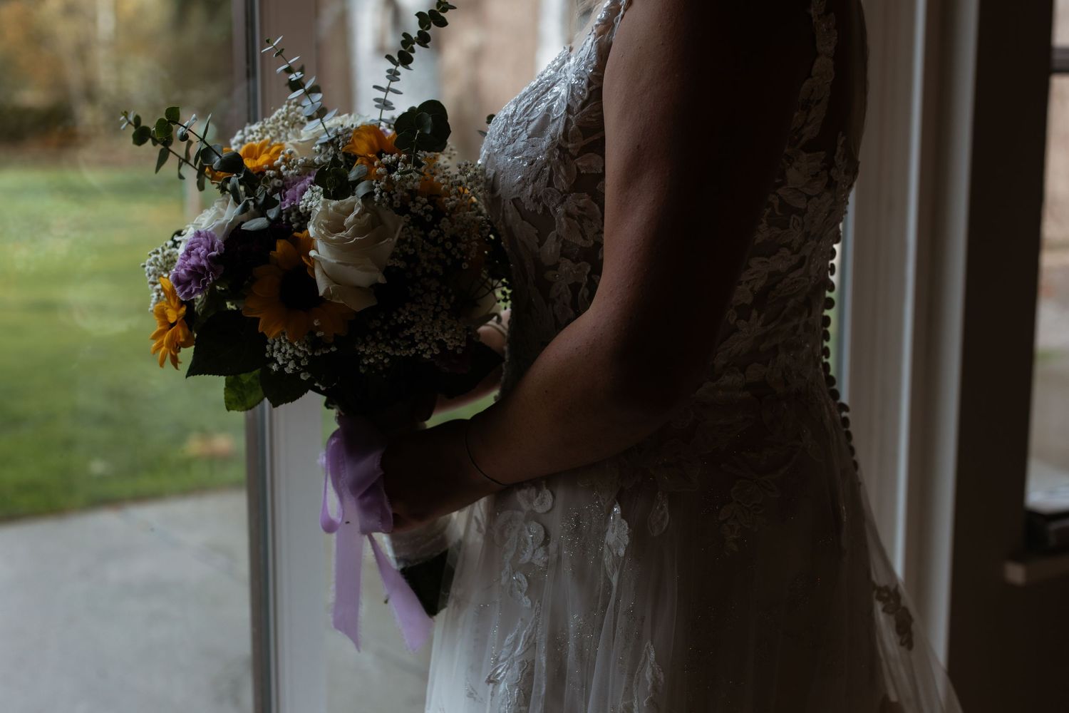 Close-up of bride holding rustic bouquet with sunflowers and white roses tied with lavender ribbon.