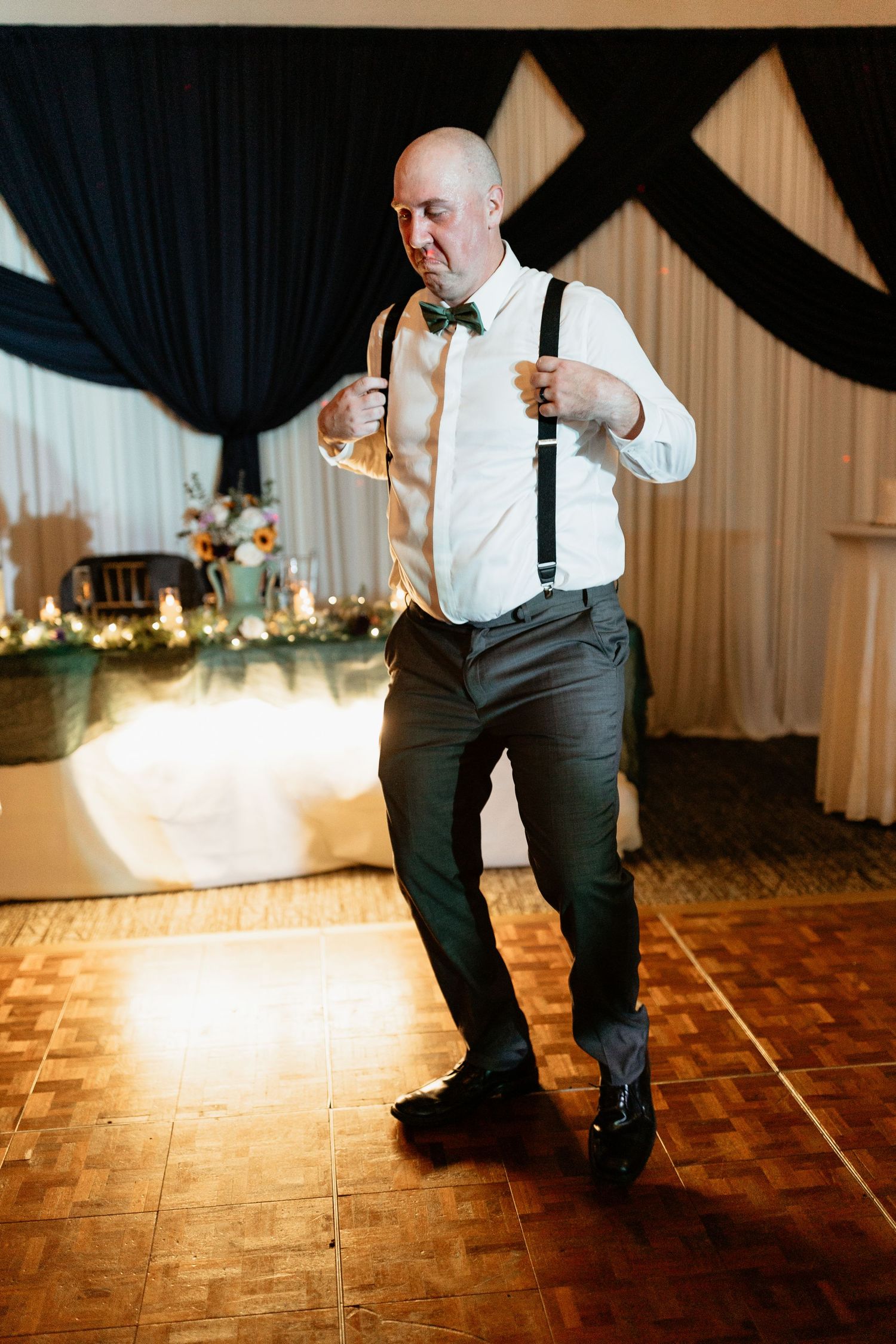 Bald man in white shirt, suspenders, and green bow tie dancing at a wedding reception on a wooden dance floor.