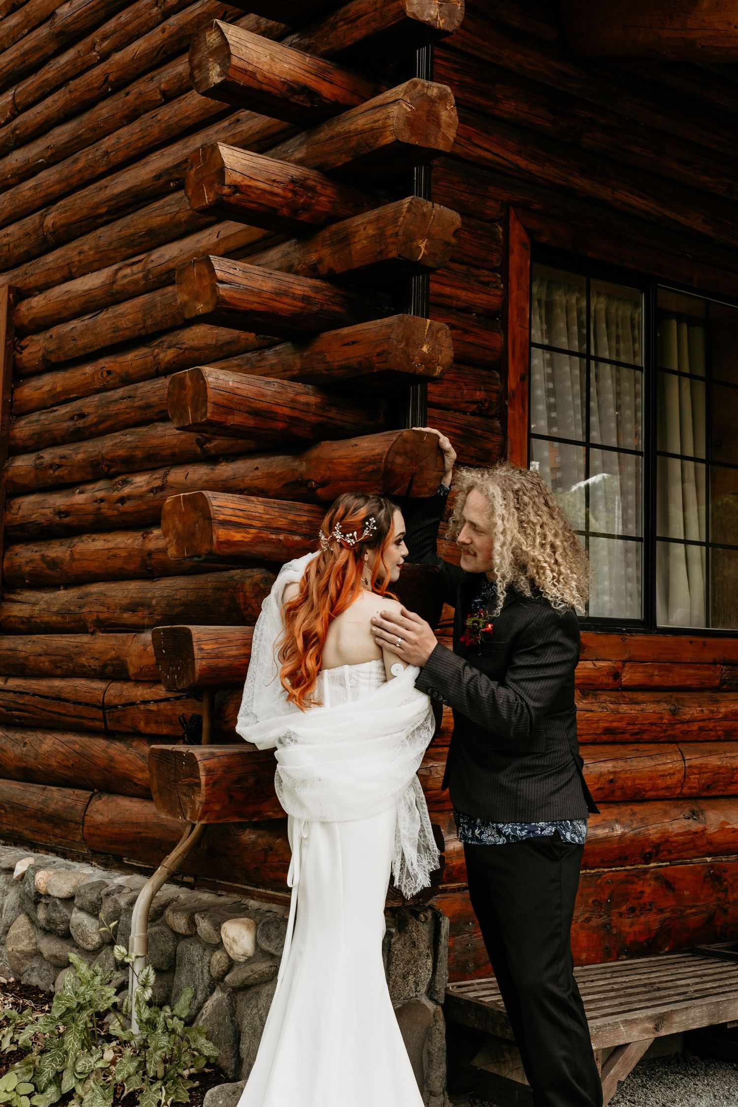 Bride in white gown and groom in black suit share a tender moment outside a rustic log cabin wedding venue.