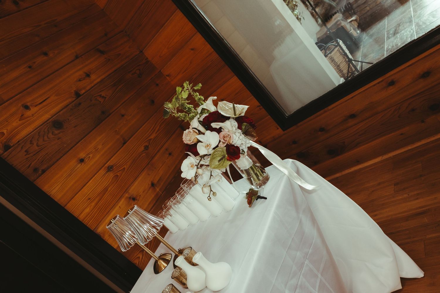 Elegant wedding table with white linen, floral arrangement of burgundy and white blooms on rustic wood floor.