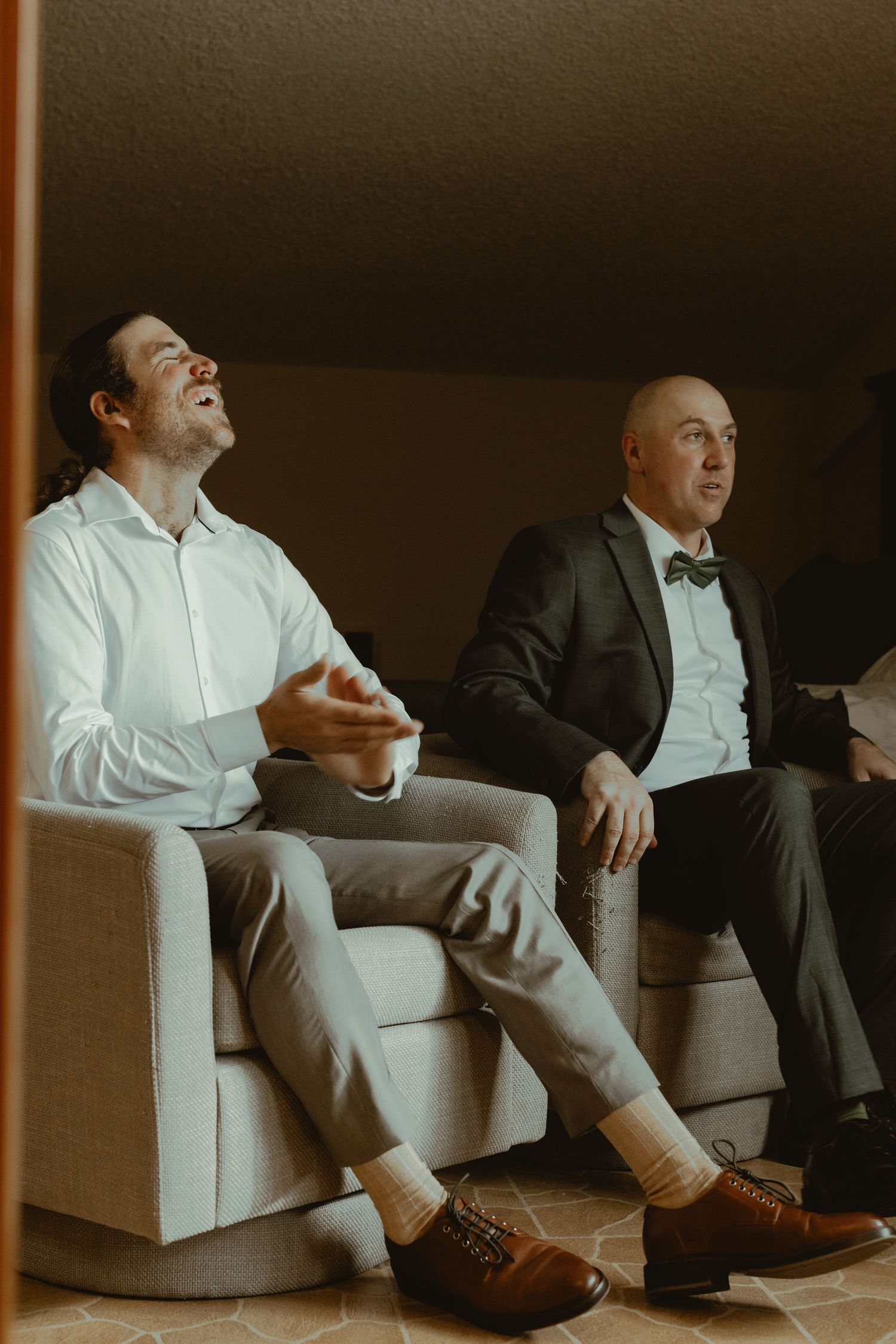Two groomsmen relax and laugh together on a sofa while getting ready before the wedding ceremony.