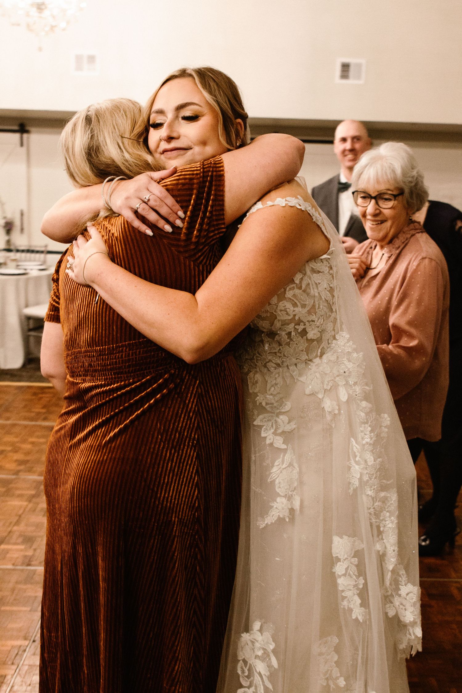 Bride in lace wedding gown shares a heartfelt hug with a guest in a rust velvet dress.