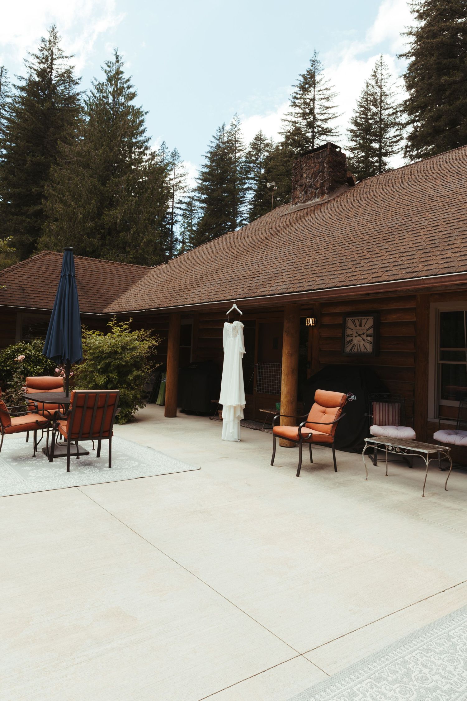 Wedding dress hanging outside a rustic log cabin patio surrounded by tall pine trees with outdoor seating area.