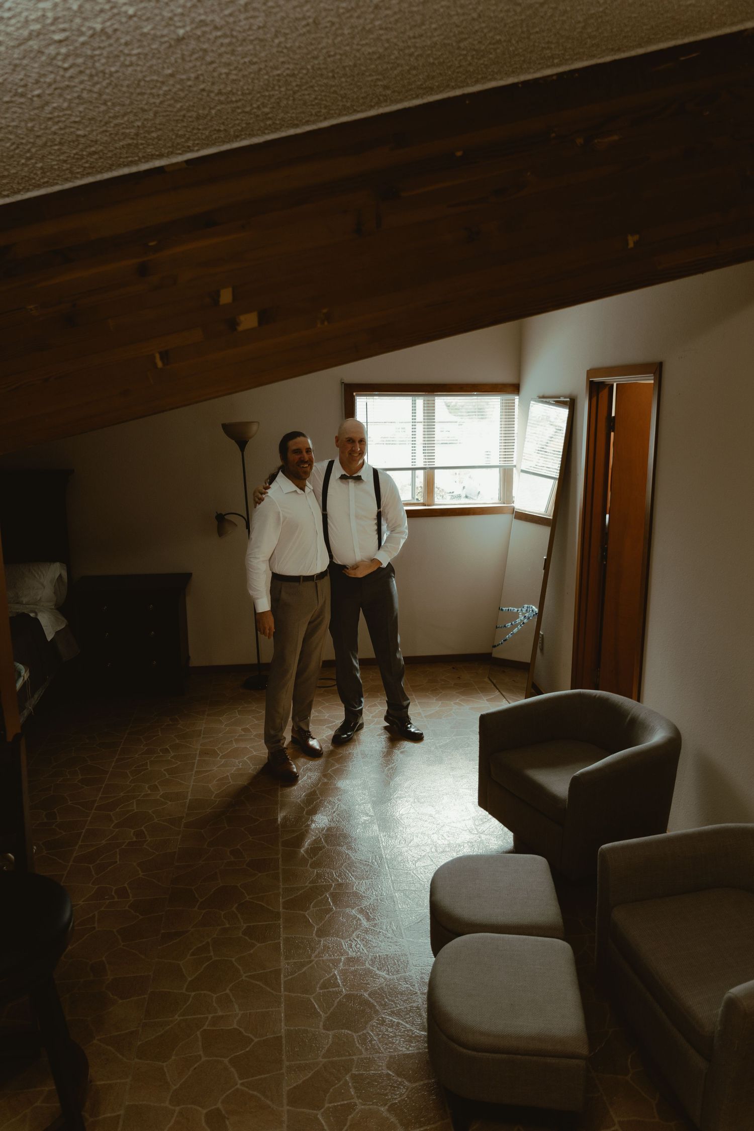 Two men in white shirts and suspenders pose together in a rustic room with wood beam ceiling.