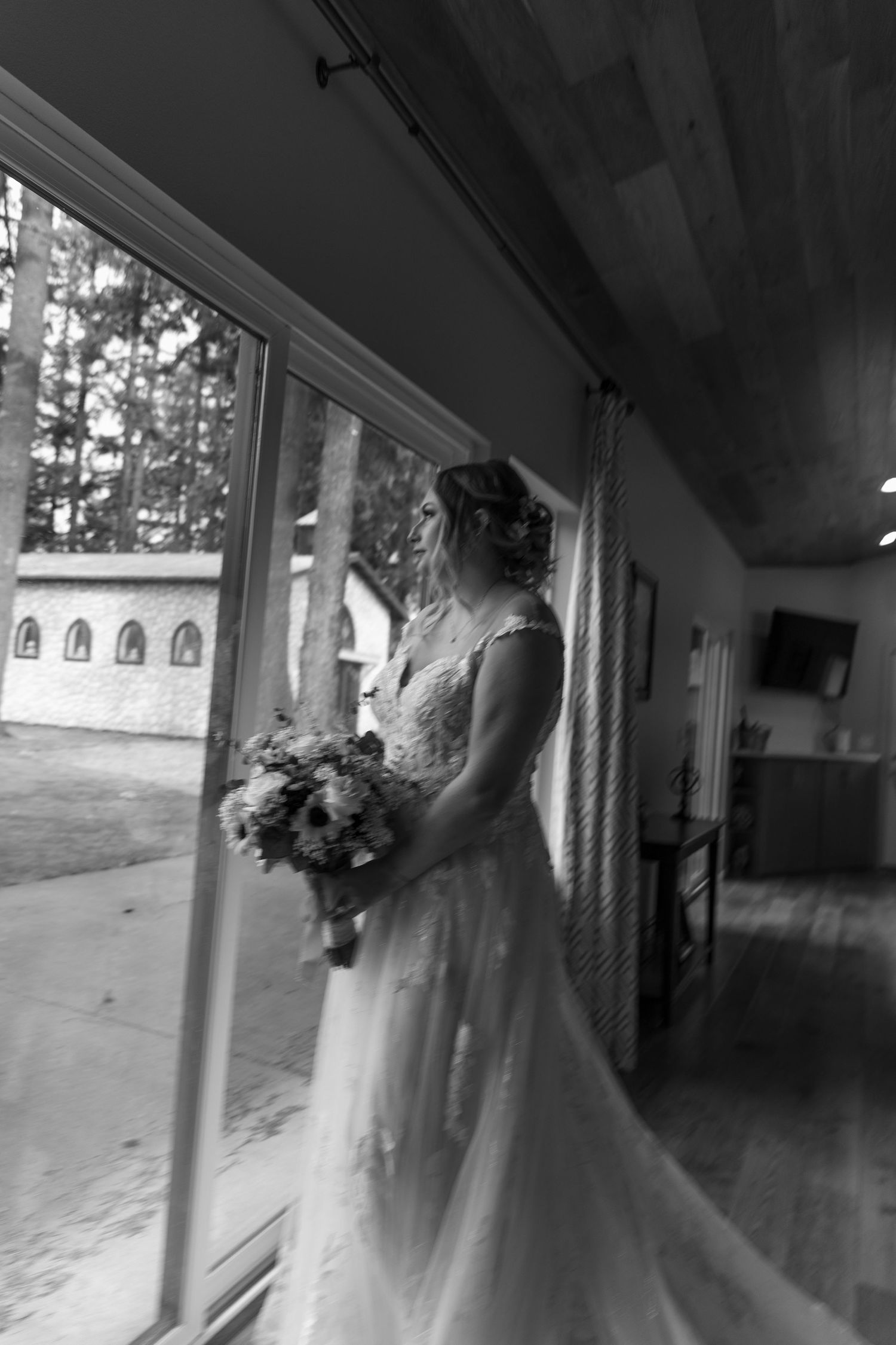 Black and white photo of bride in lace gown holding bouquet beside large window.