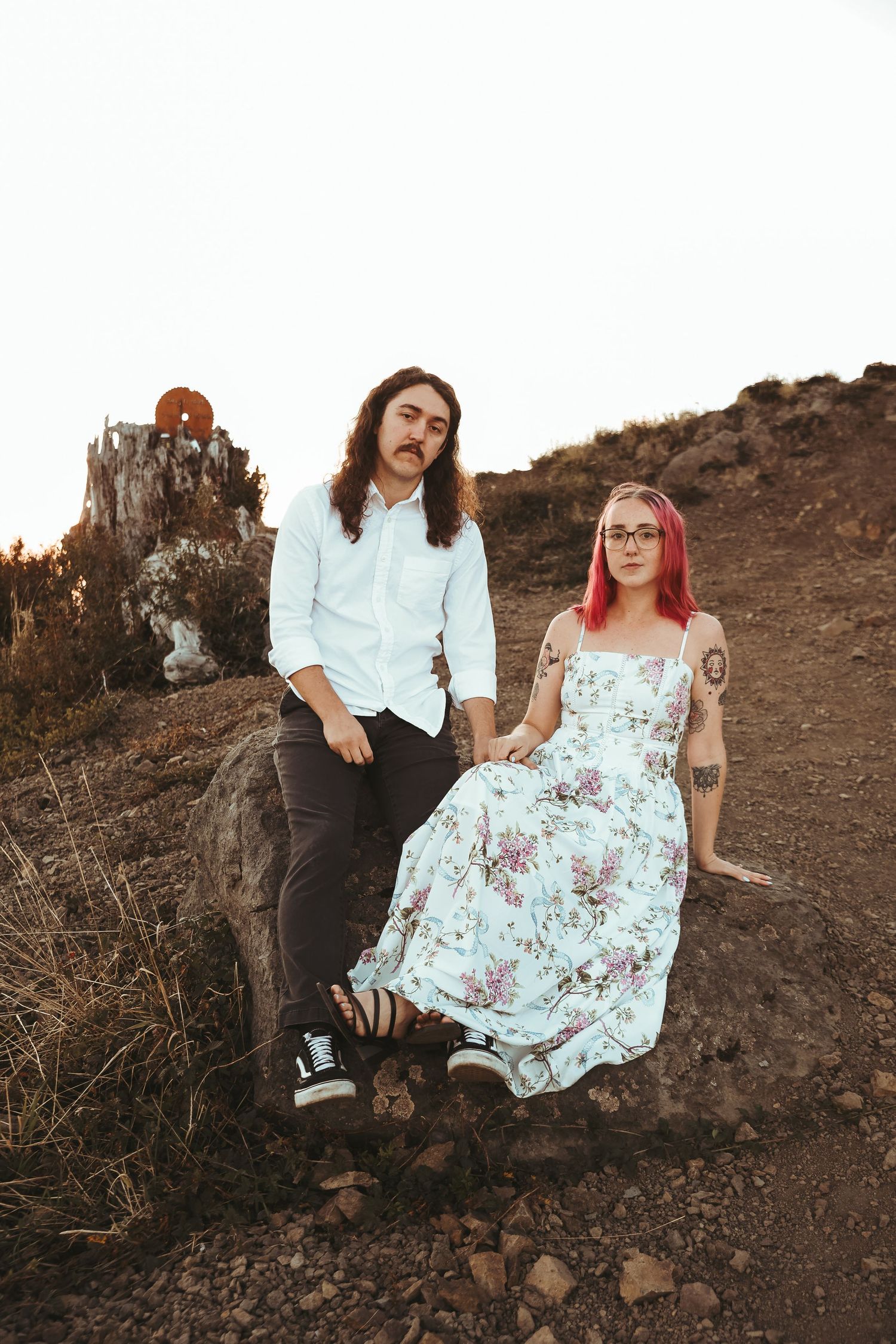A couple poses outdoors on rocky terrain, man in white shirt and woman in floral dress with pink hair and tattoos.