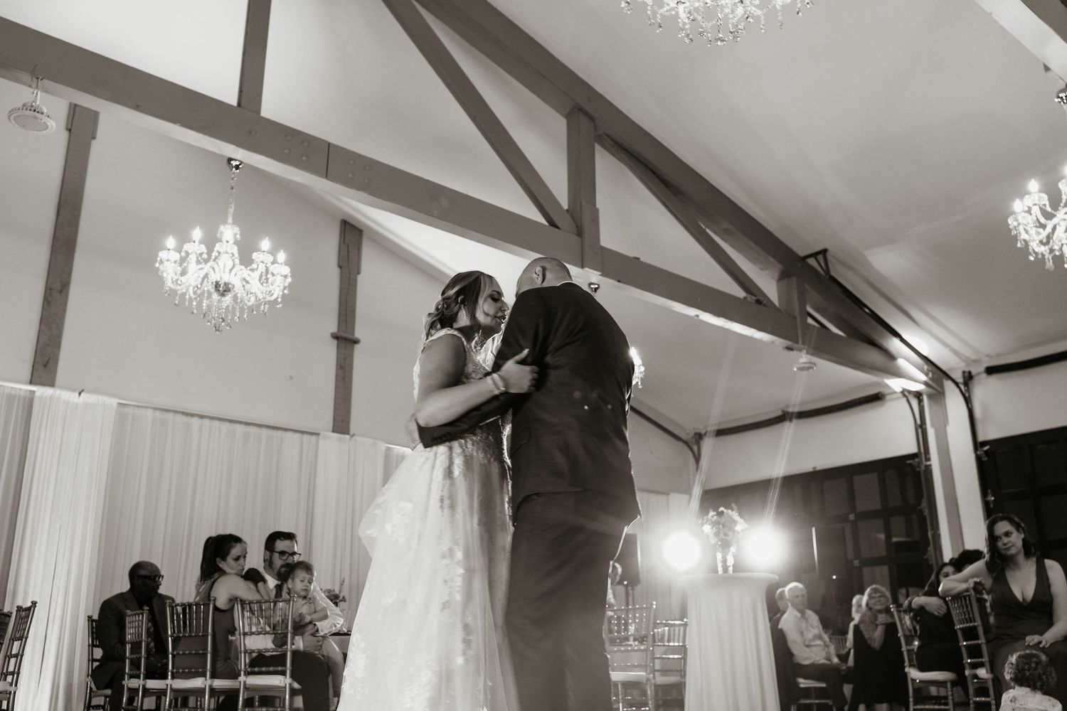 Bride and groom share an emotional first dance under chandeliers in a rustic wedding venue.