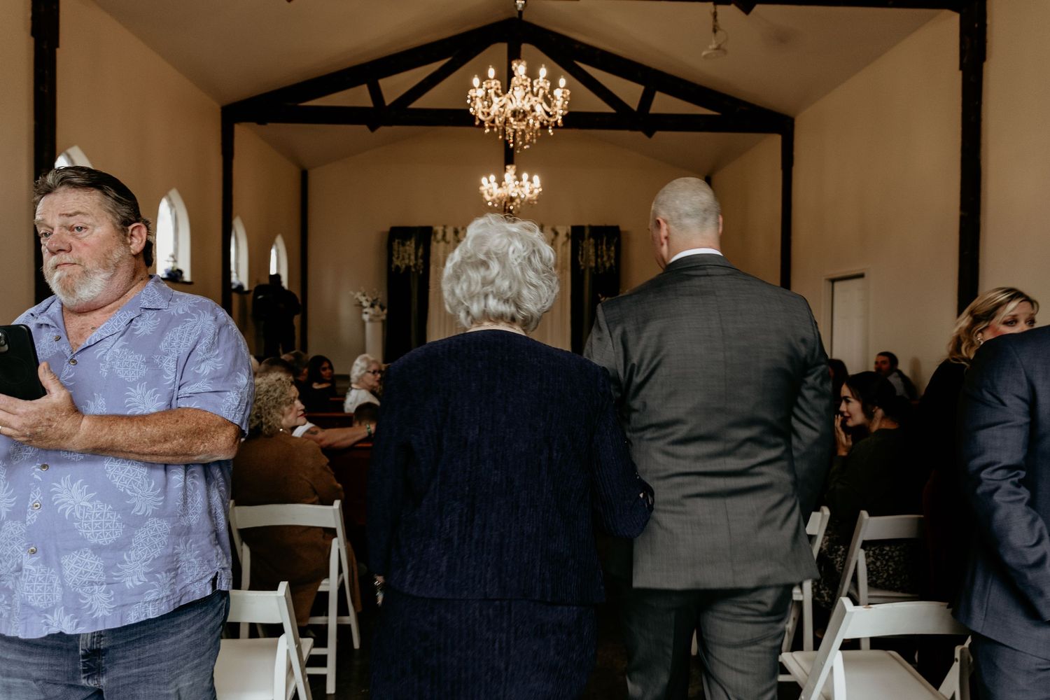 Wedding guests gathered inside a rustic chapel with a chandelier before the ceremony begins.