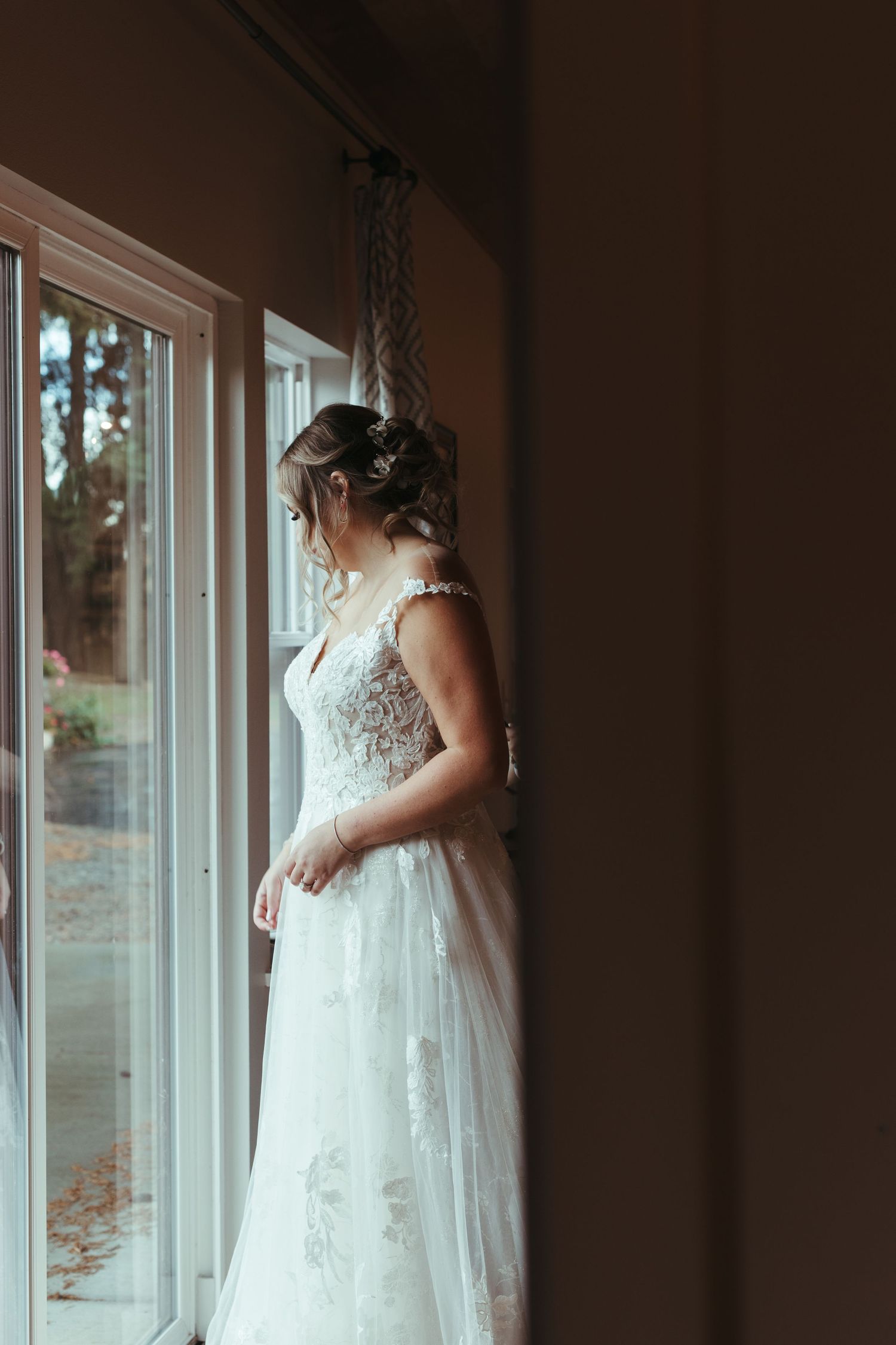 Bride in elegant lace wedding gown gazing out large window before her ceremony.
