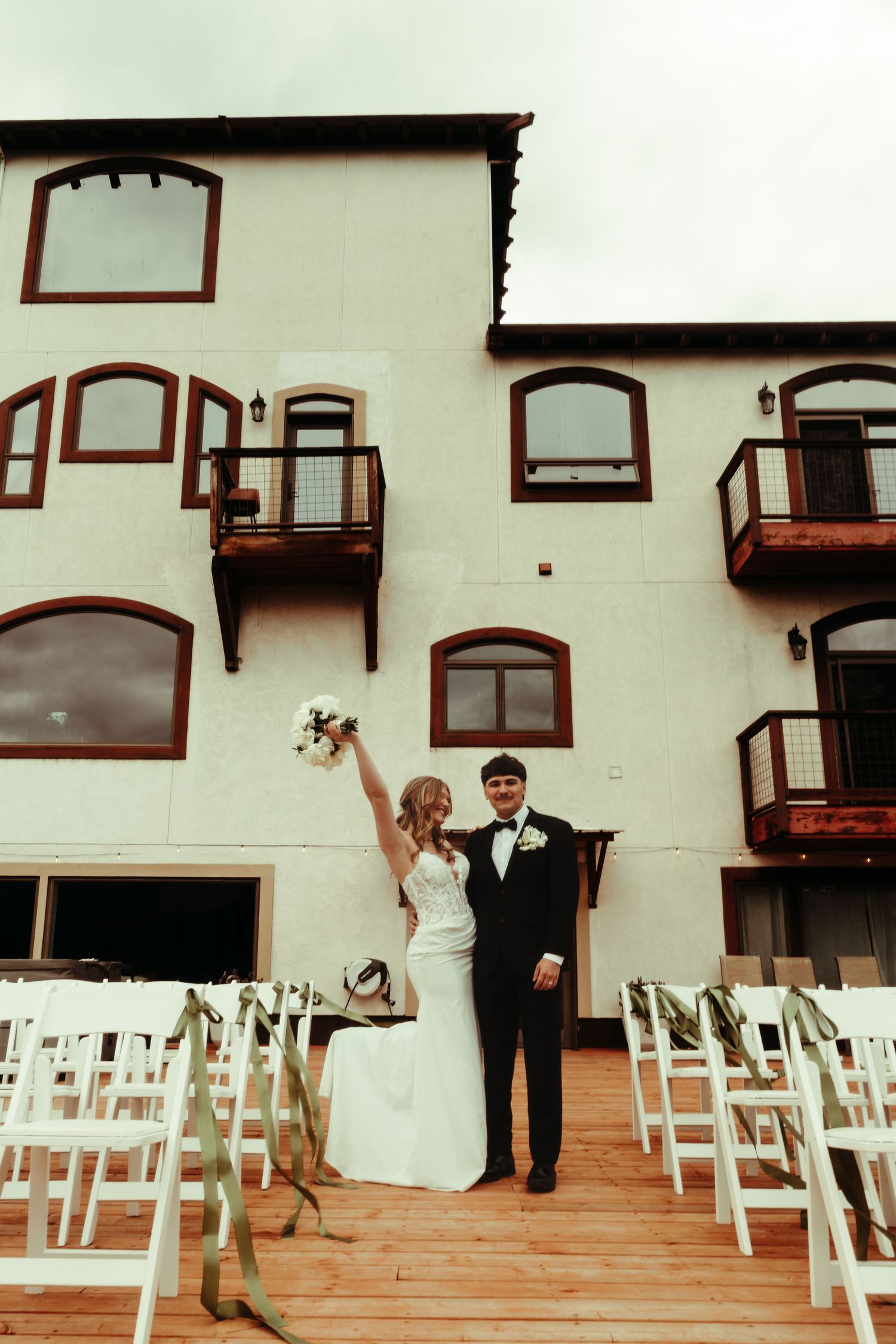 Bride raises bouquet joyfully beside groom in tuxedo on wooden deck outside white wedding venue with balconies.