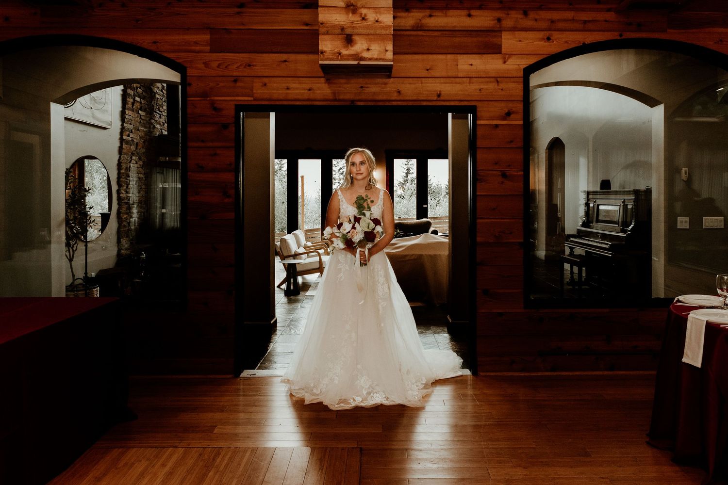 Bride in white gown holding bouquet stands in rustic wood-paneled venue with arched doorways and warm lighting.