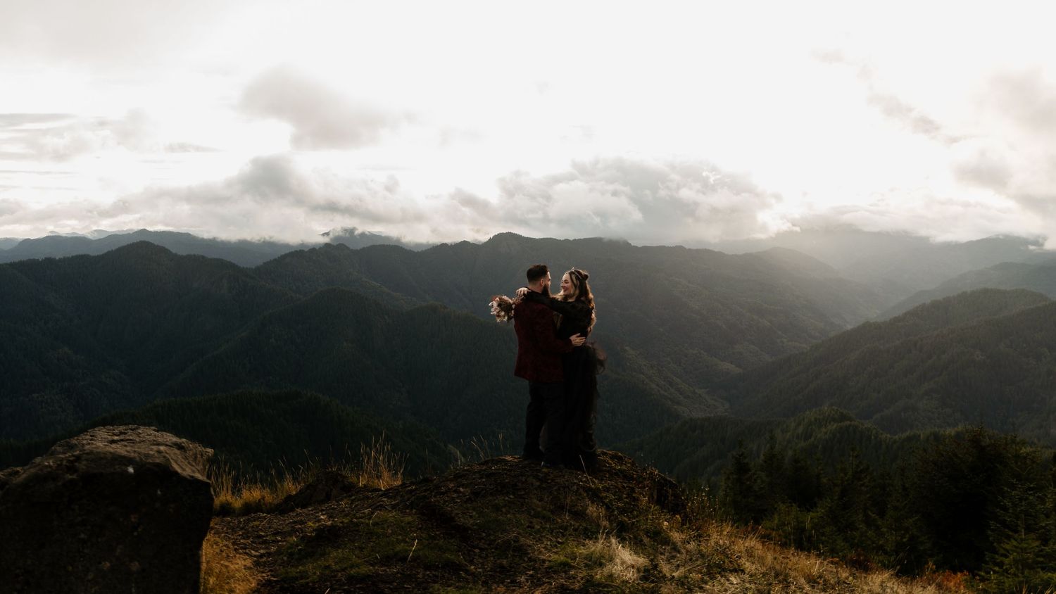Couple embracing on a rocky mountaintop overlooking breathtaking misty mountain ranges at sunset.
