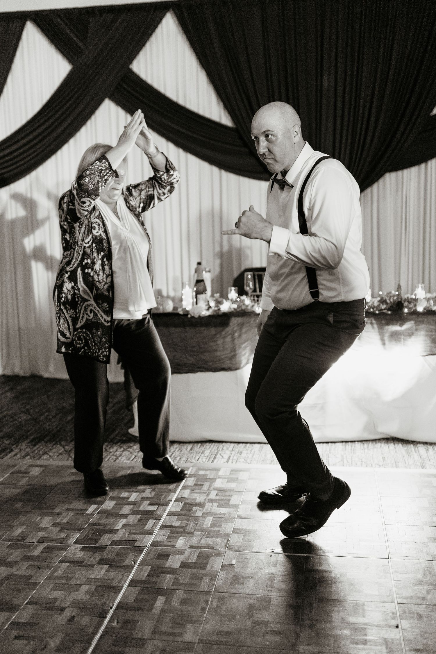 Black and white photo of a bald man in suspenders and bow tie dancing energetically with an older woman.