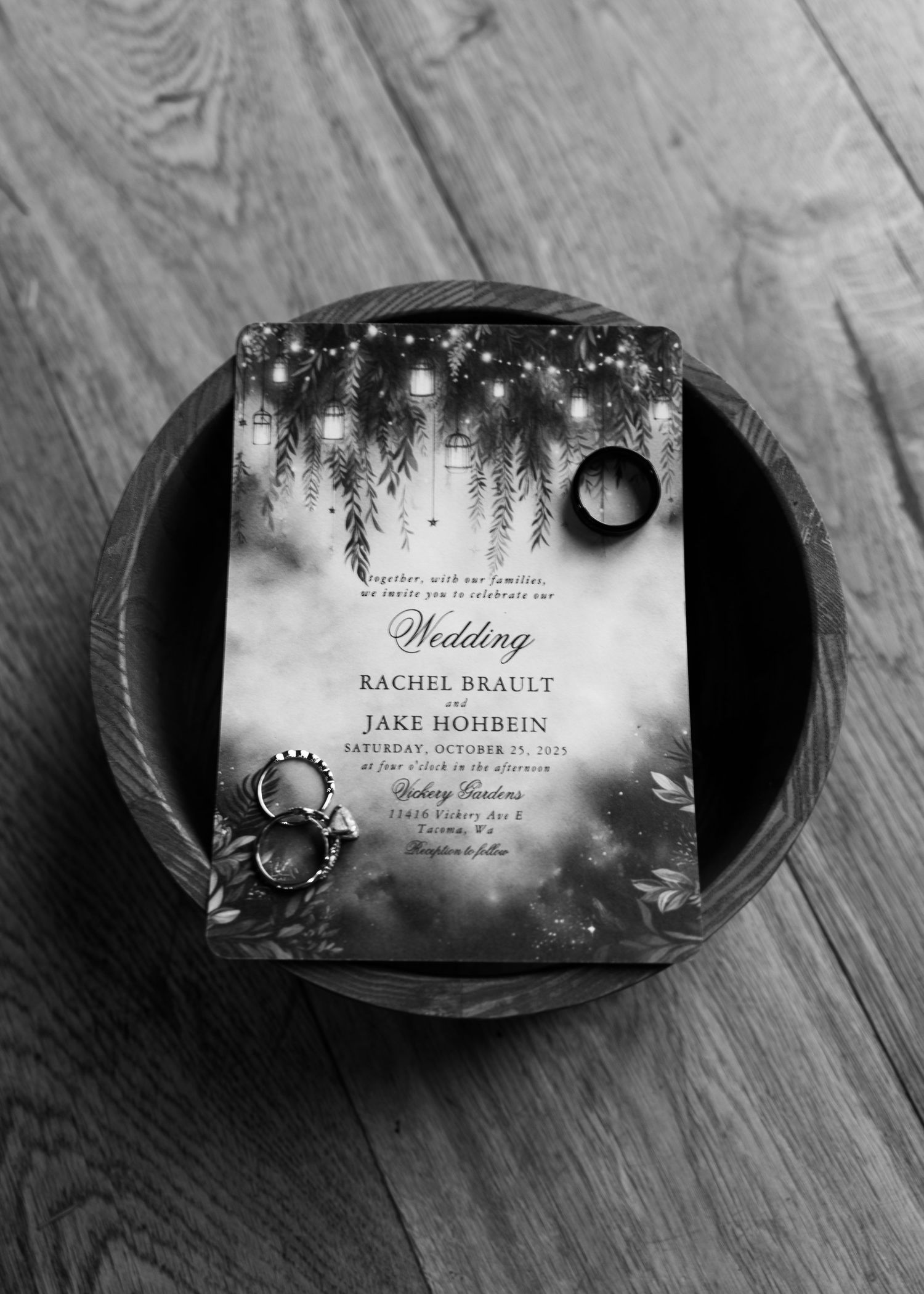 Black and white flat lay of enchanted forest wedding invitation with rings in a rustic wooden bowl.