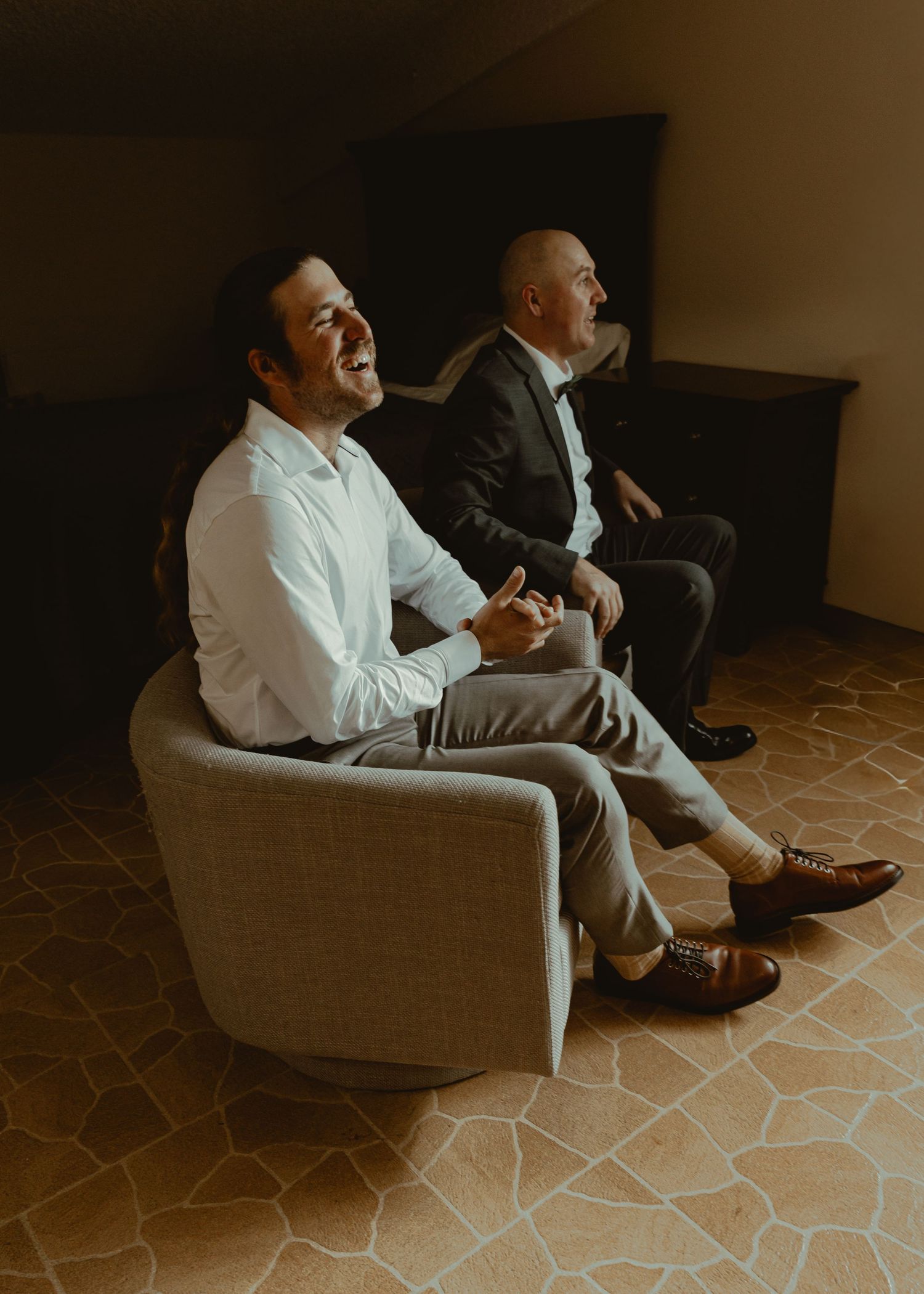 Two men in formal wedding attire laugh and relax together while seated in a warmly lit room.