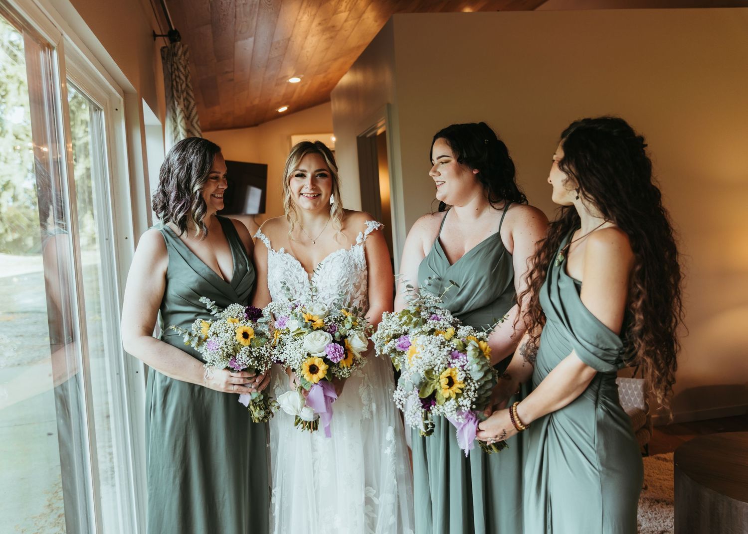 Bride in lace gown stands with three bridesmaids in sage green dresses holding colorful bouquets by a window.