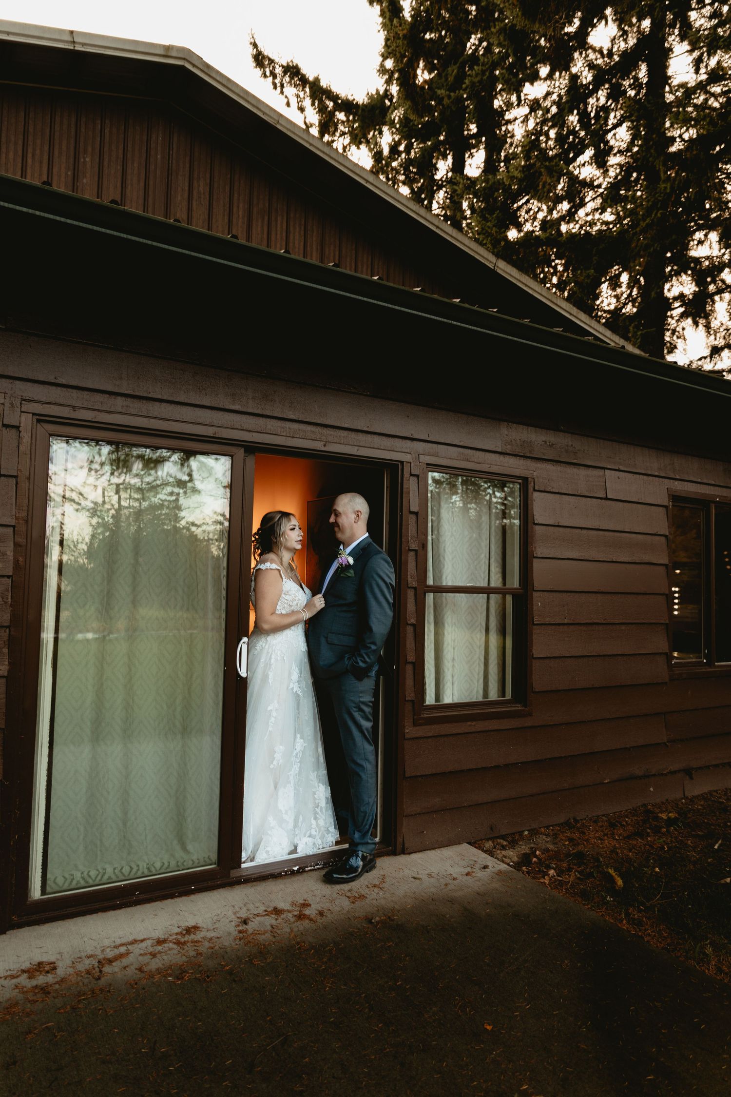 Bride and groom standing in doorway of rustic brown building sharing a romantic moment at sunset.