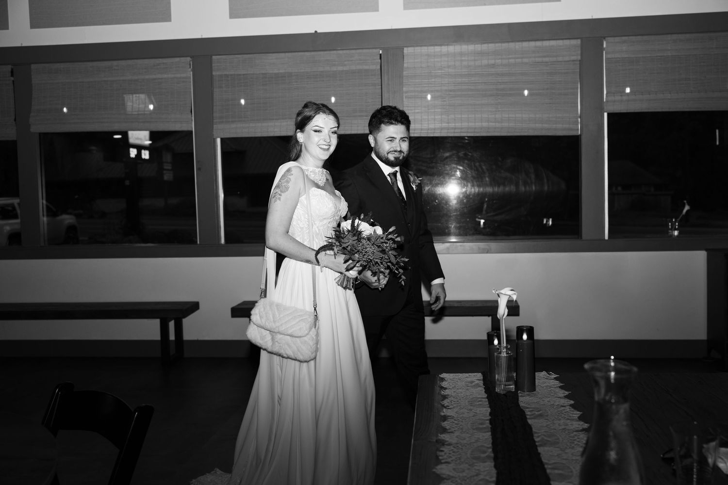 Black and white photo of a bride in a long gown holding a bouquet standing beside a groom in a dark suit at a wedding reception.