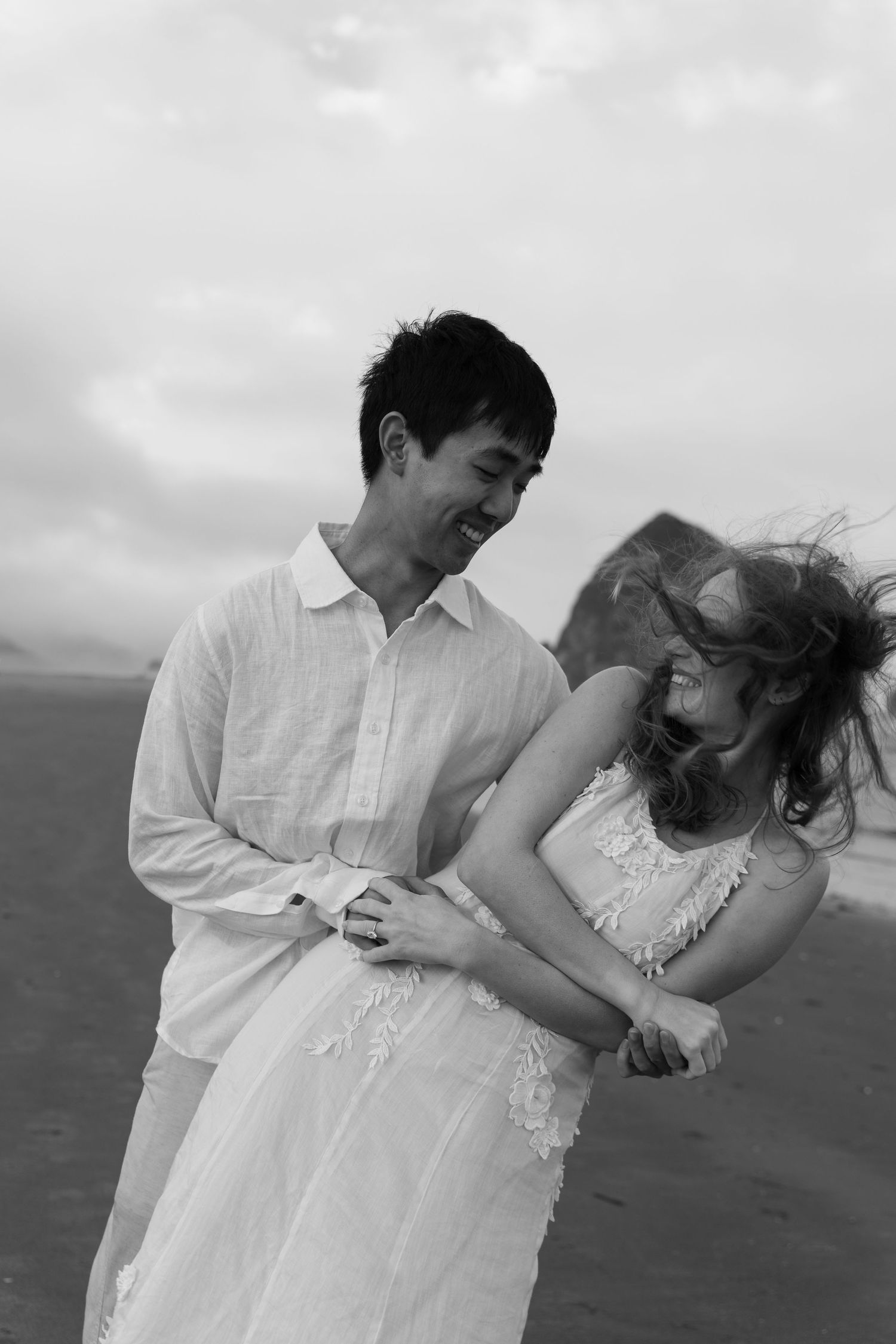 Black and white photo of a smiling couple embracing on a windy beach, wearing white outfits with ocean in background.
