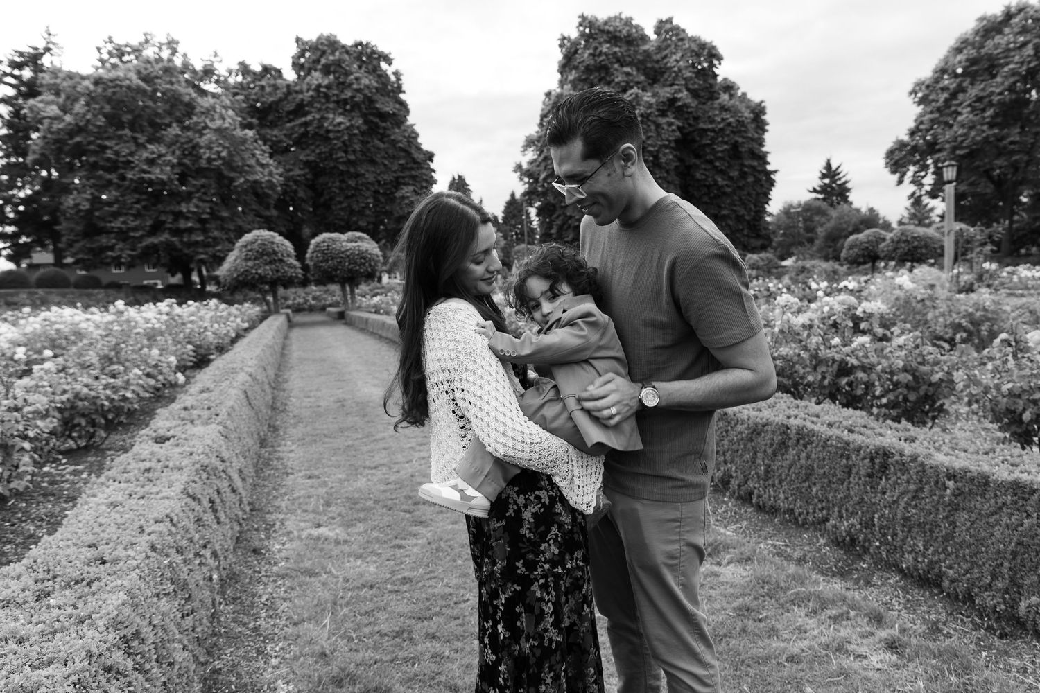 Black and white photo of a couple admiring their baby in a lush garden setting.