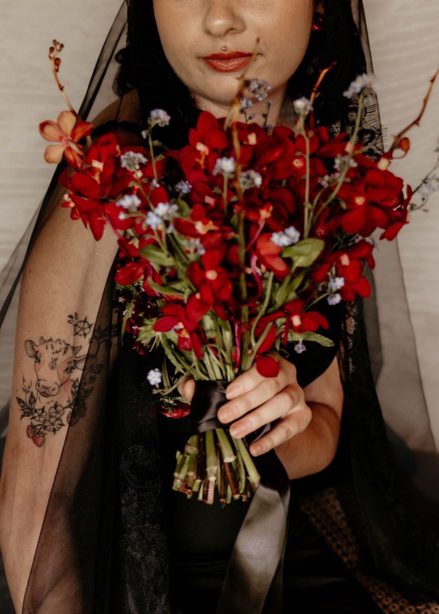 Bride in black lace veil holds red floral bouquet with tattoo visible on arm.