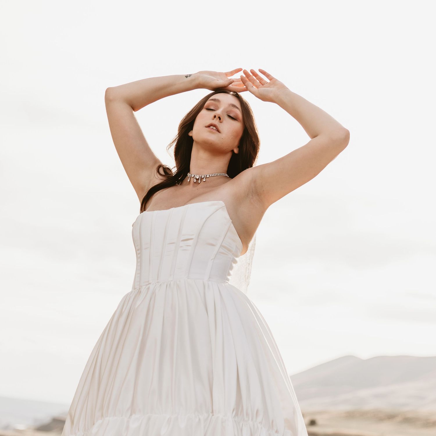 Woman in a white strapless corset-style gown with a full pleated skirt posing with raised arms against a soft, hazy desert landscape.