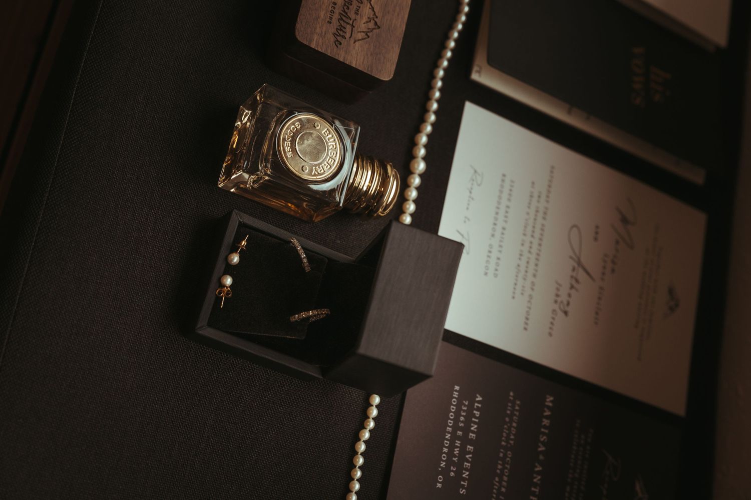Wedding flat lay with a perfume bottle, open ring box with gold bands, pearl strand, and black and gold invitation cards.
