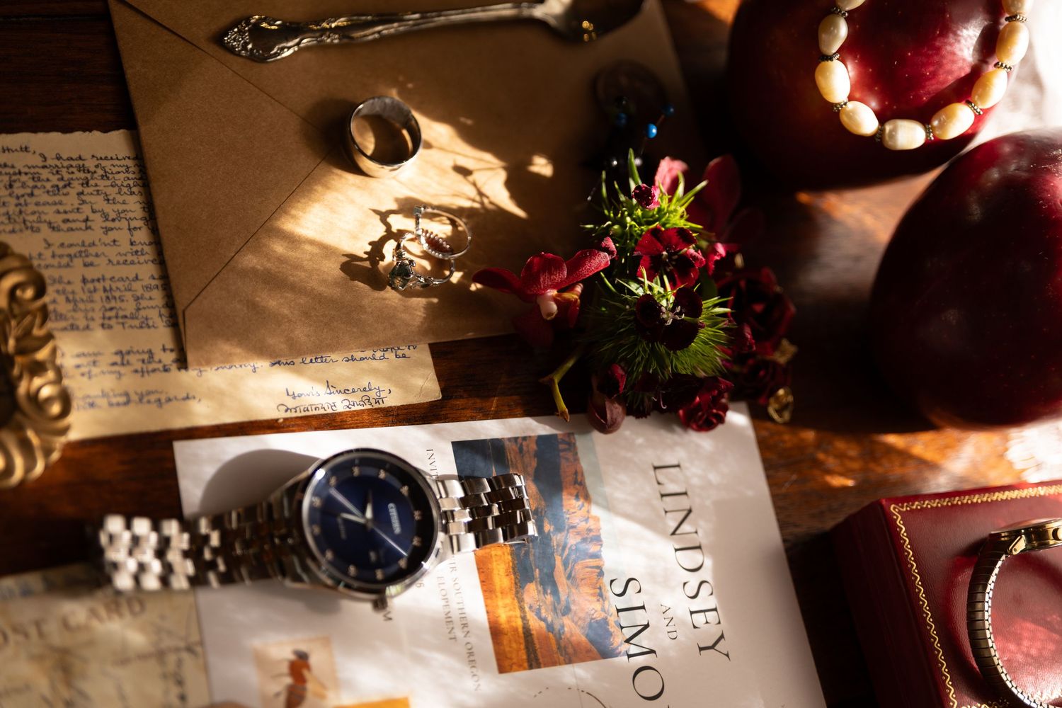Flat lay of a silver watch, red flowers, handwritten letters, and jewelry on a wooden table in warm sunlight.