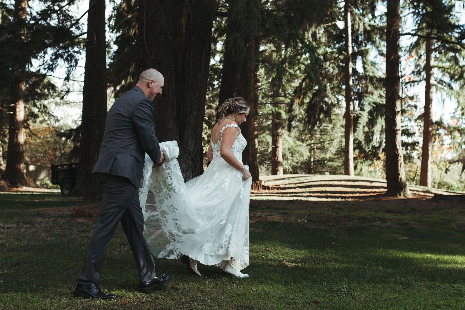 Groom in gray suit helping bride carry her lace wedding dress train across a green lawn outdoors.