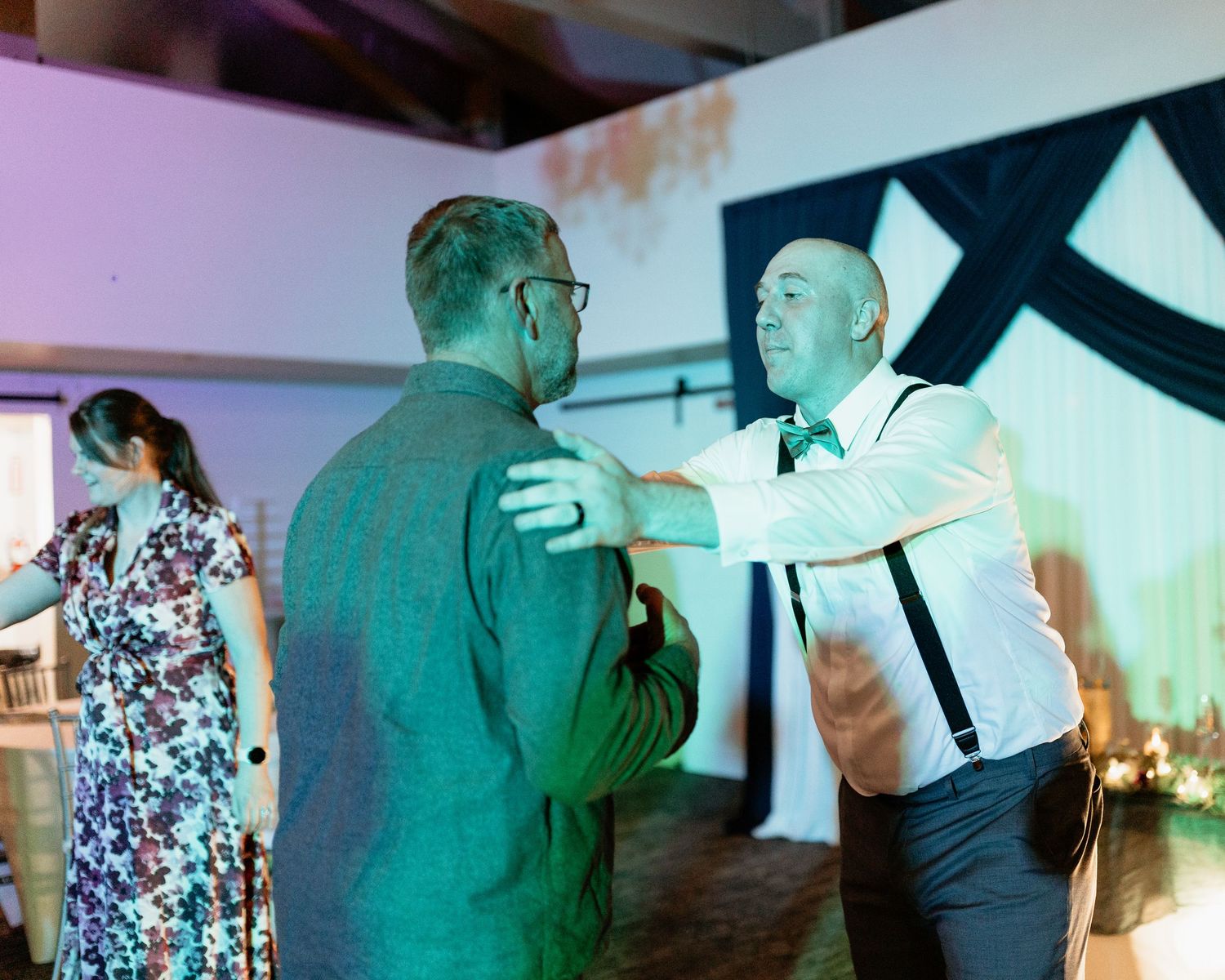 Two men share a warm embrace on a wedding reception dance floor under green ambient lighting.