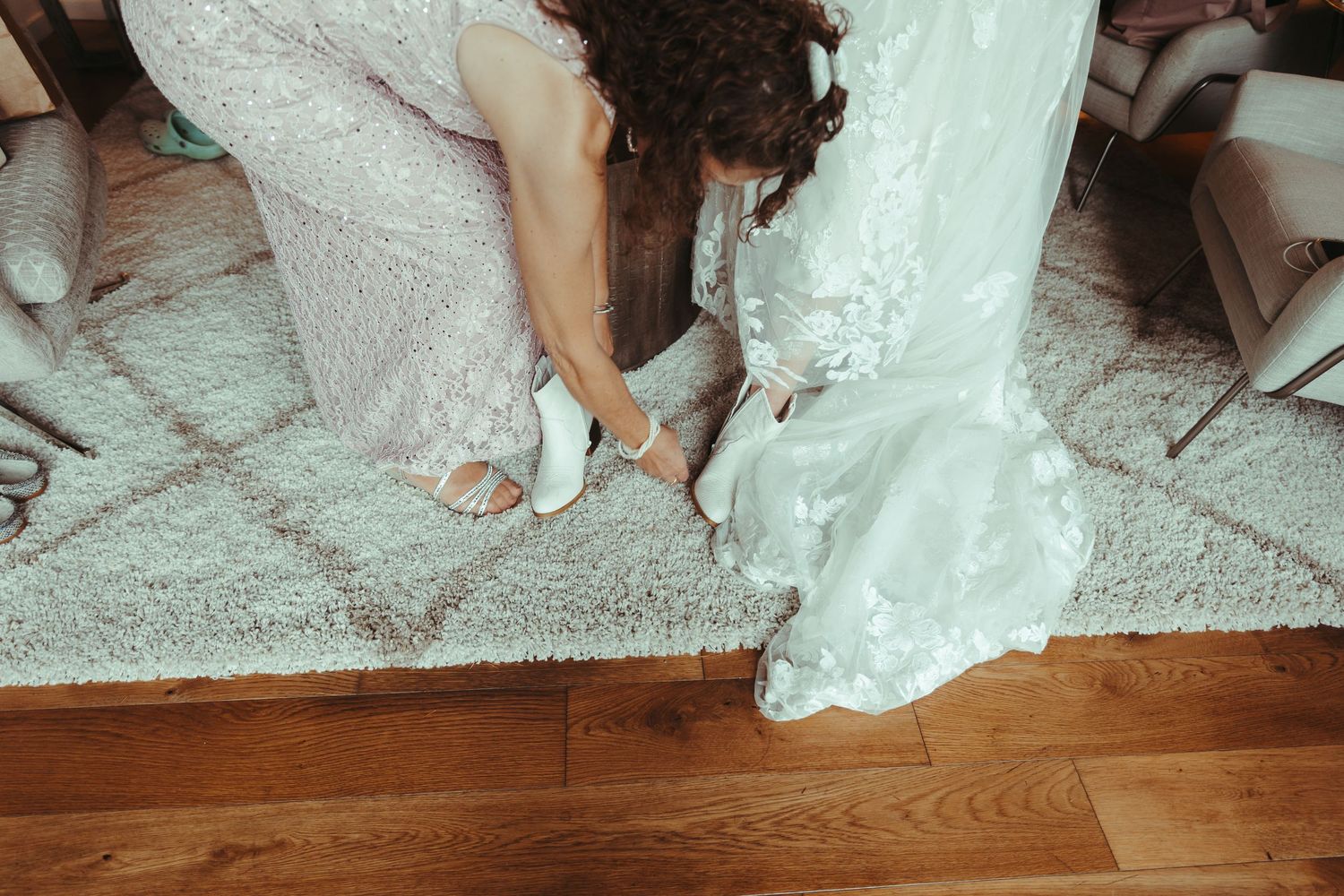 Helper adjusting white bridal heels on bride in lace wedding gown on a patterned rug.