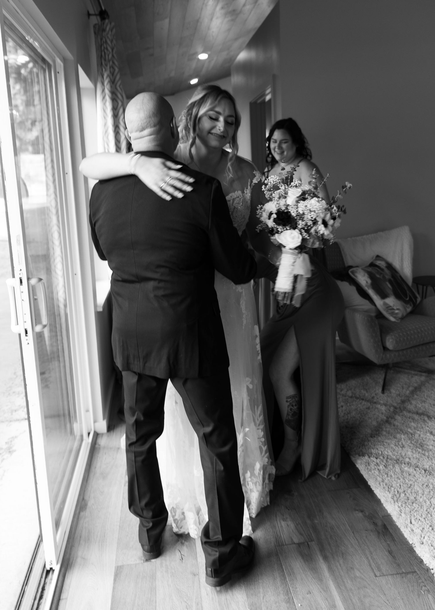 Emotional black and white photo of bride in wedding gown embracing a man while bridesmaid watches nearby.