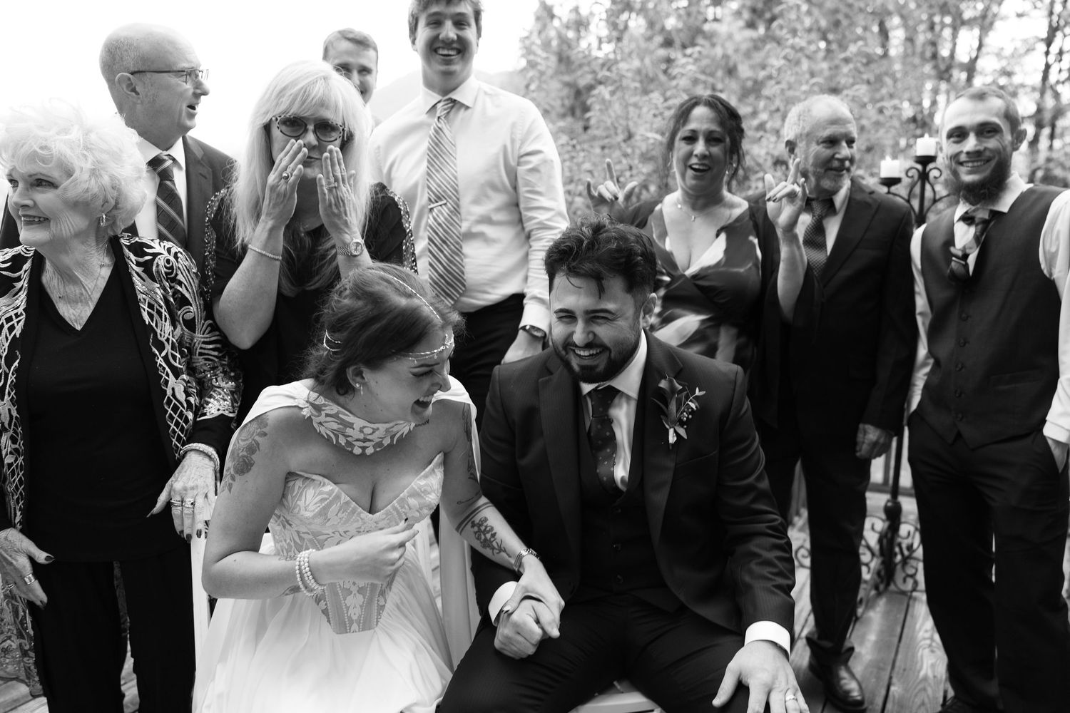 Joyful wedding guests laugh and cheer around a smiling bride and groom seated together during a lively ceremony moment in black and white.