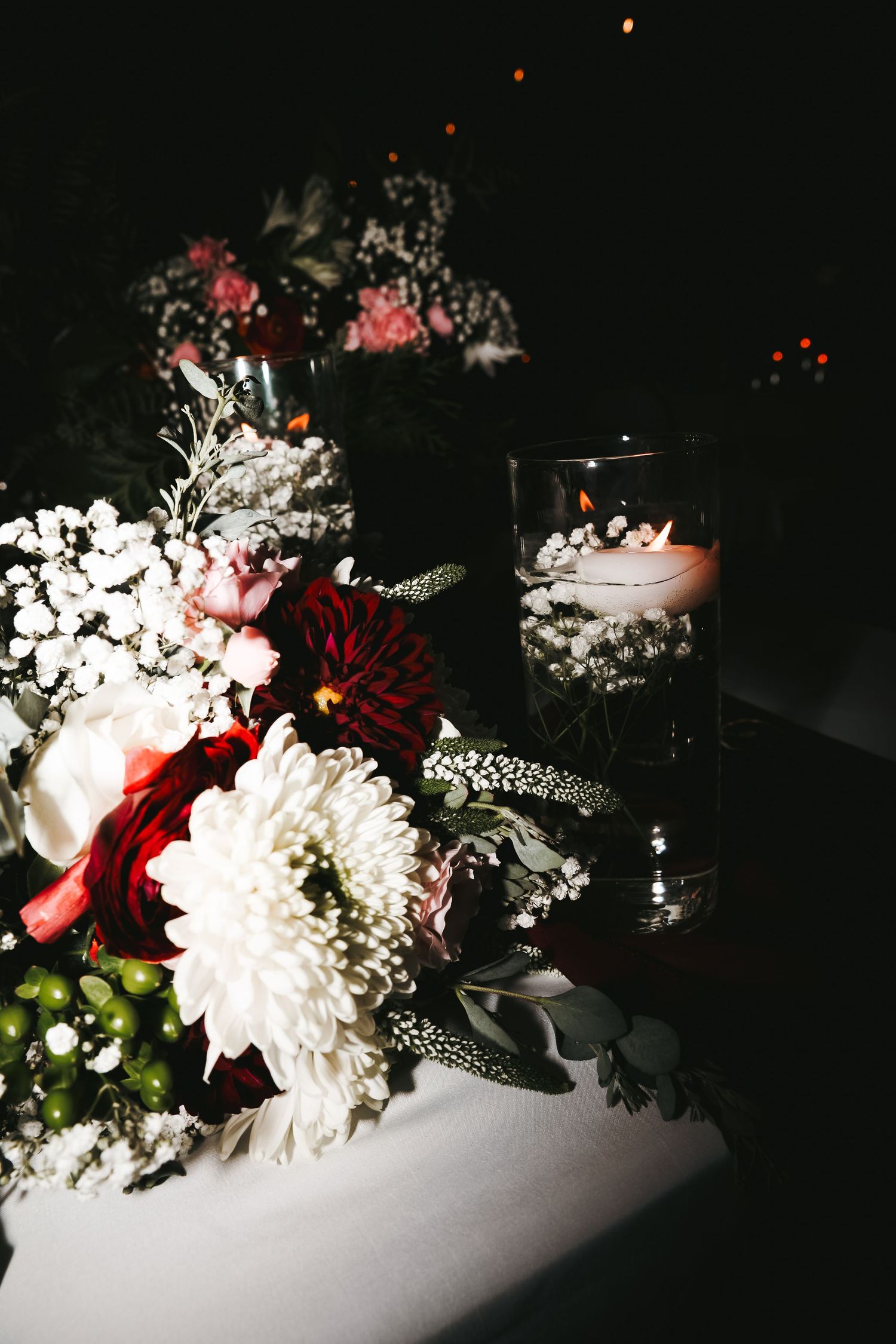Wedding table centerpiece with burgundy dahlias, white chrysanthemums, baby's breath and a floating candle in a glass vase at night.