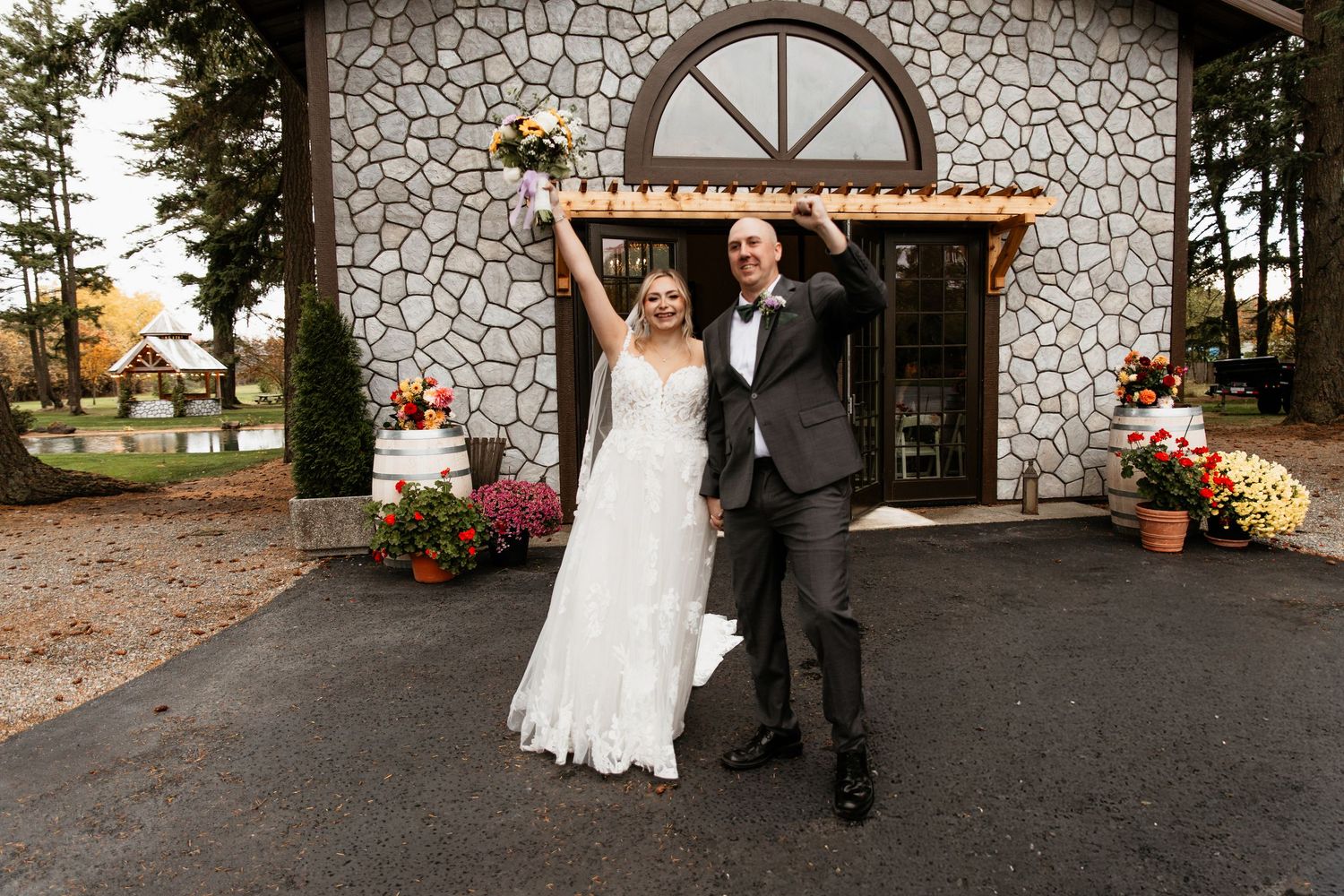 Joyful newlyweds celebrate outside a stone chapel surrounded by autumn flowers raising their hands high.