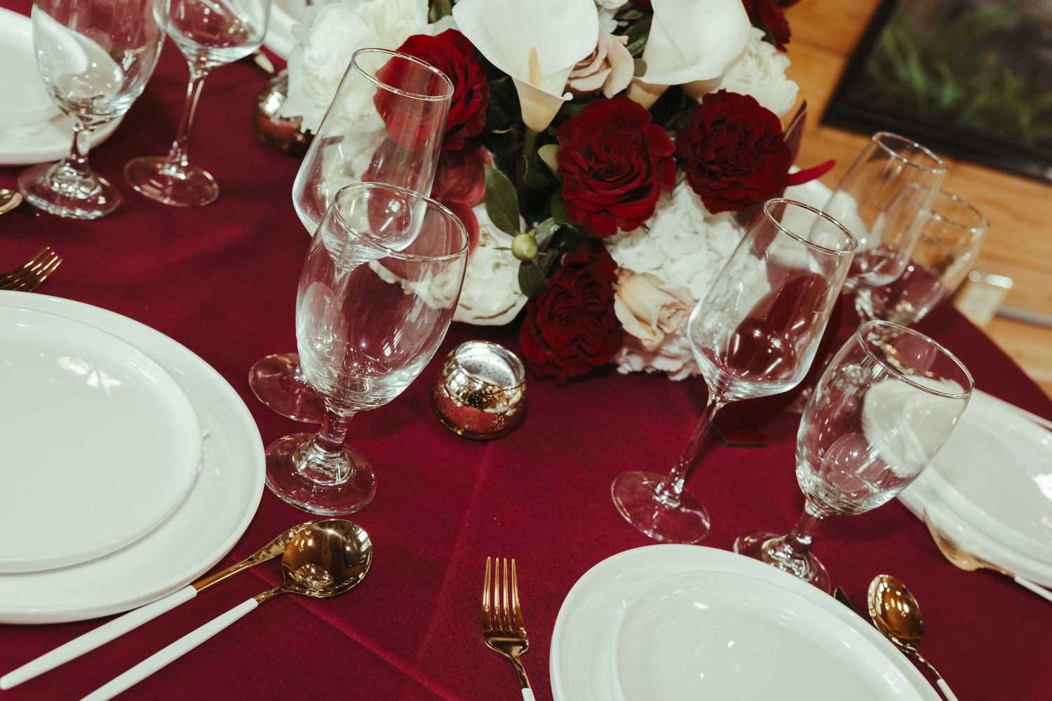 Elegant wedding table setting with burgundy linen, gold cutlery, crystal glasses, and red and white rose centerpiece.