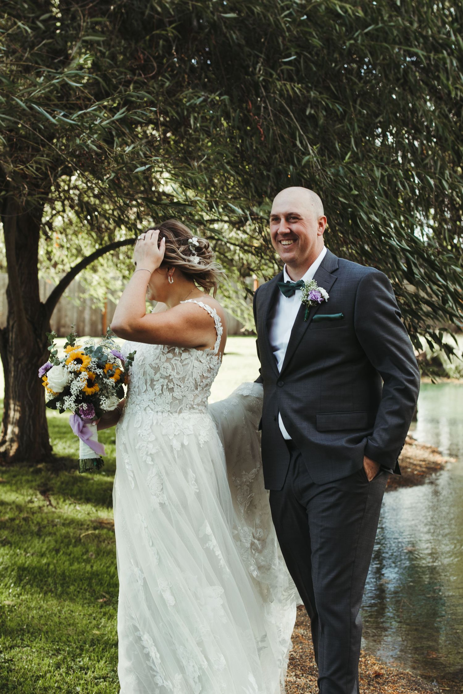 Laughing groom in blue suit with bride holding bouquet of sunflowers near a willow tree by water.