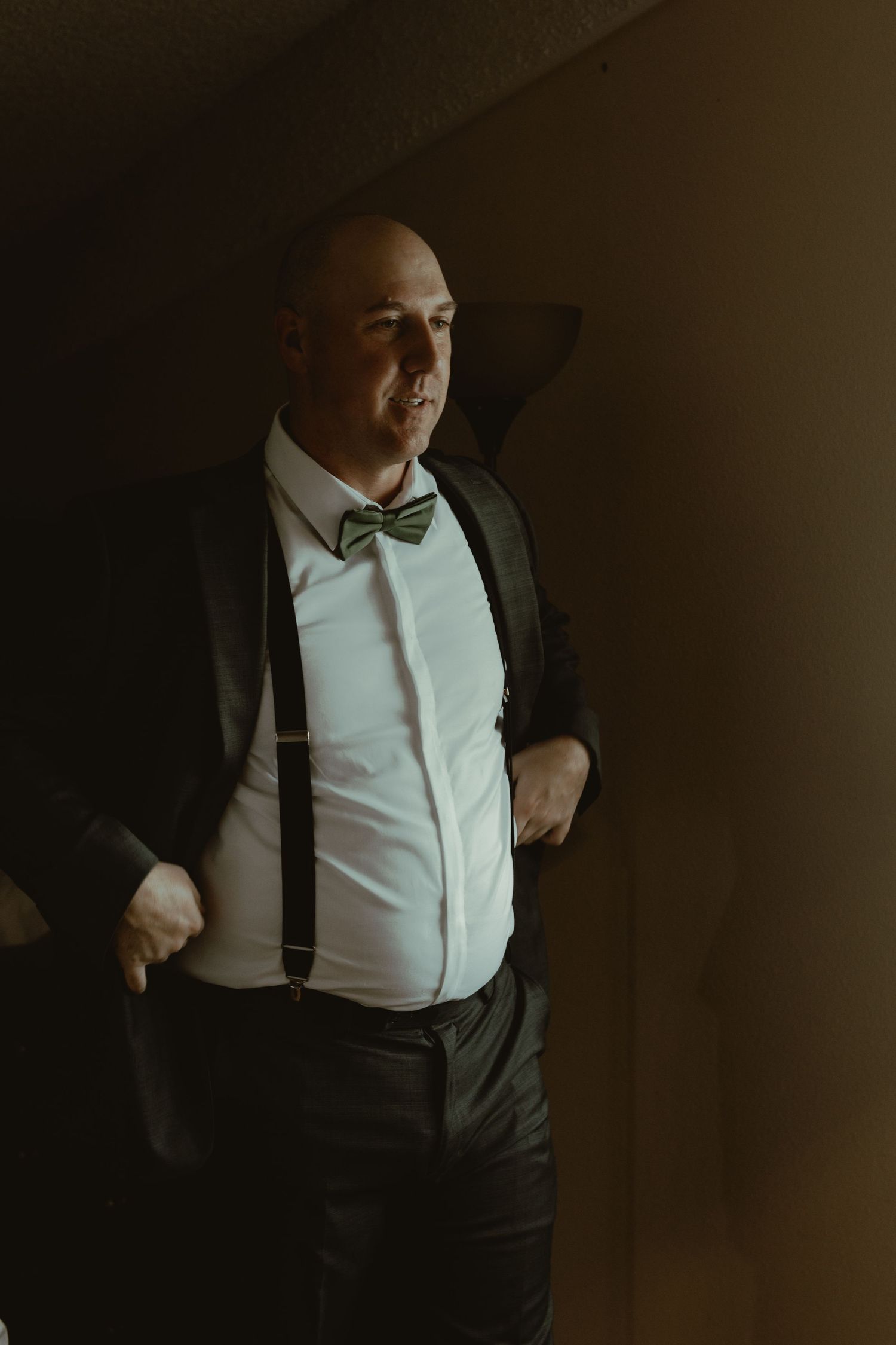 Groom in dark suit with green bow tie and suspenders poses in dramatic moody lighting before wedding.