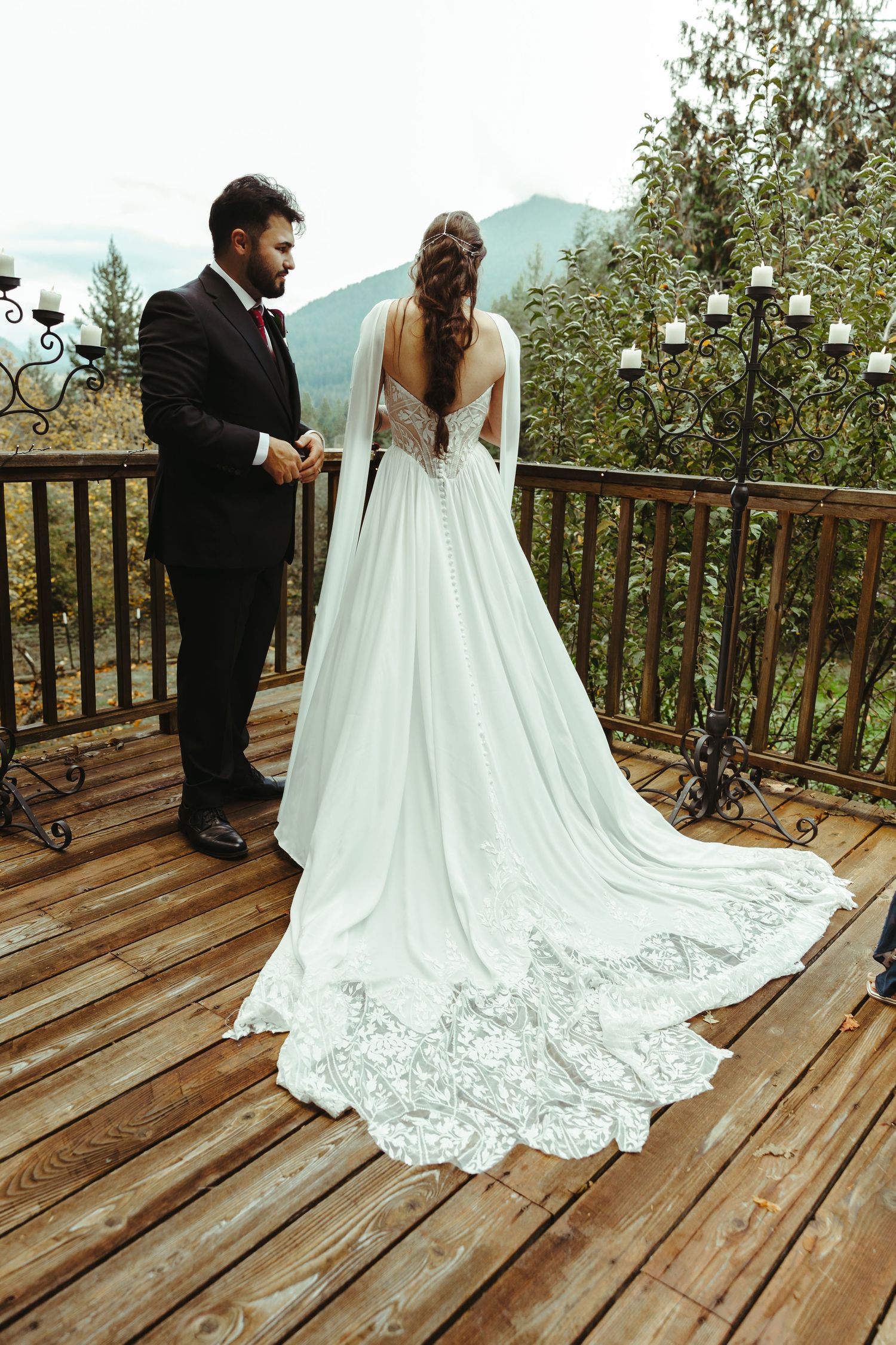 Bride in a long lace-trimmed white wedding gown with braided hair faces groom in black suit on a wooden deck with mountain views.
