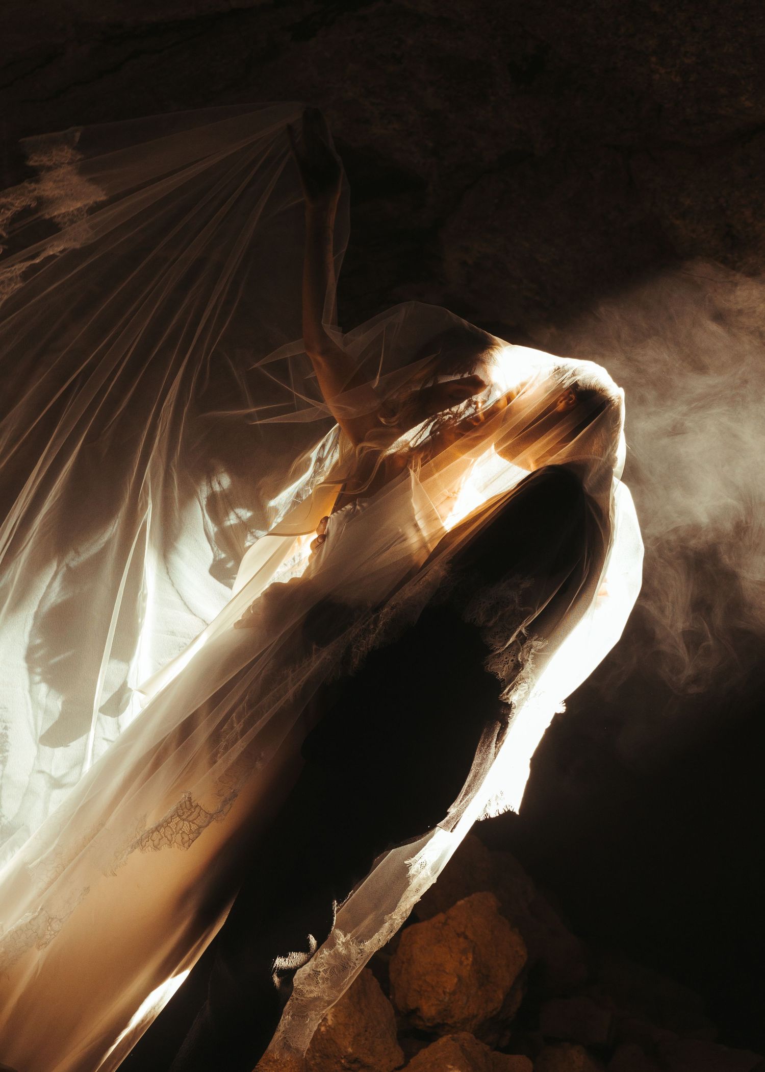 Ethereal figure draped in flowing white fabric illuminated by golden light in a dark cave setting.