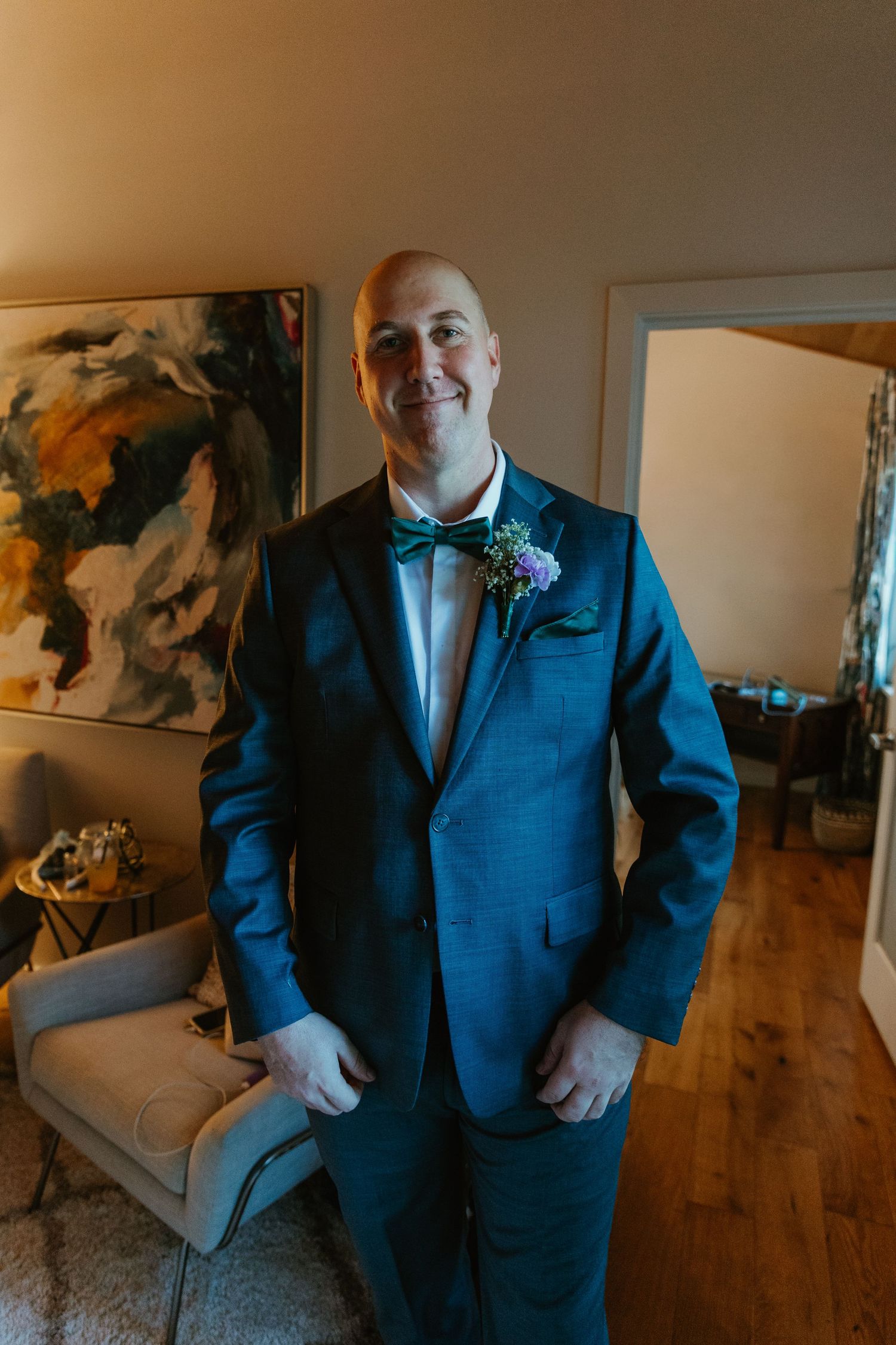 Smiling bald groom in blue suit with green bow tie and floral boutonniere posing indoors on wedding day.