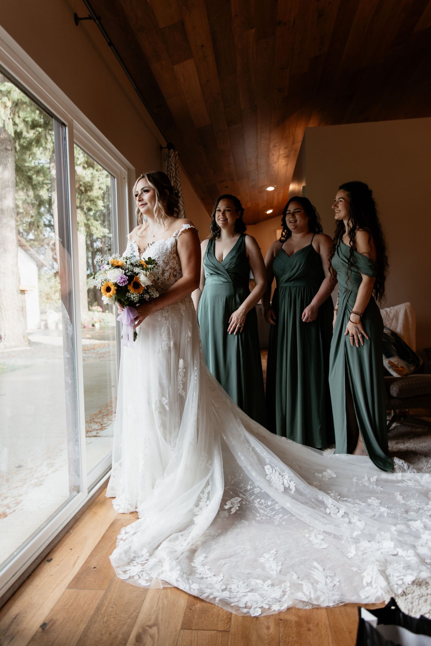 Bride with colorful bouquet standing by window with three bridesmaids in sage green gowns.