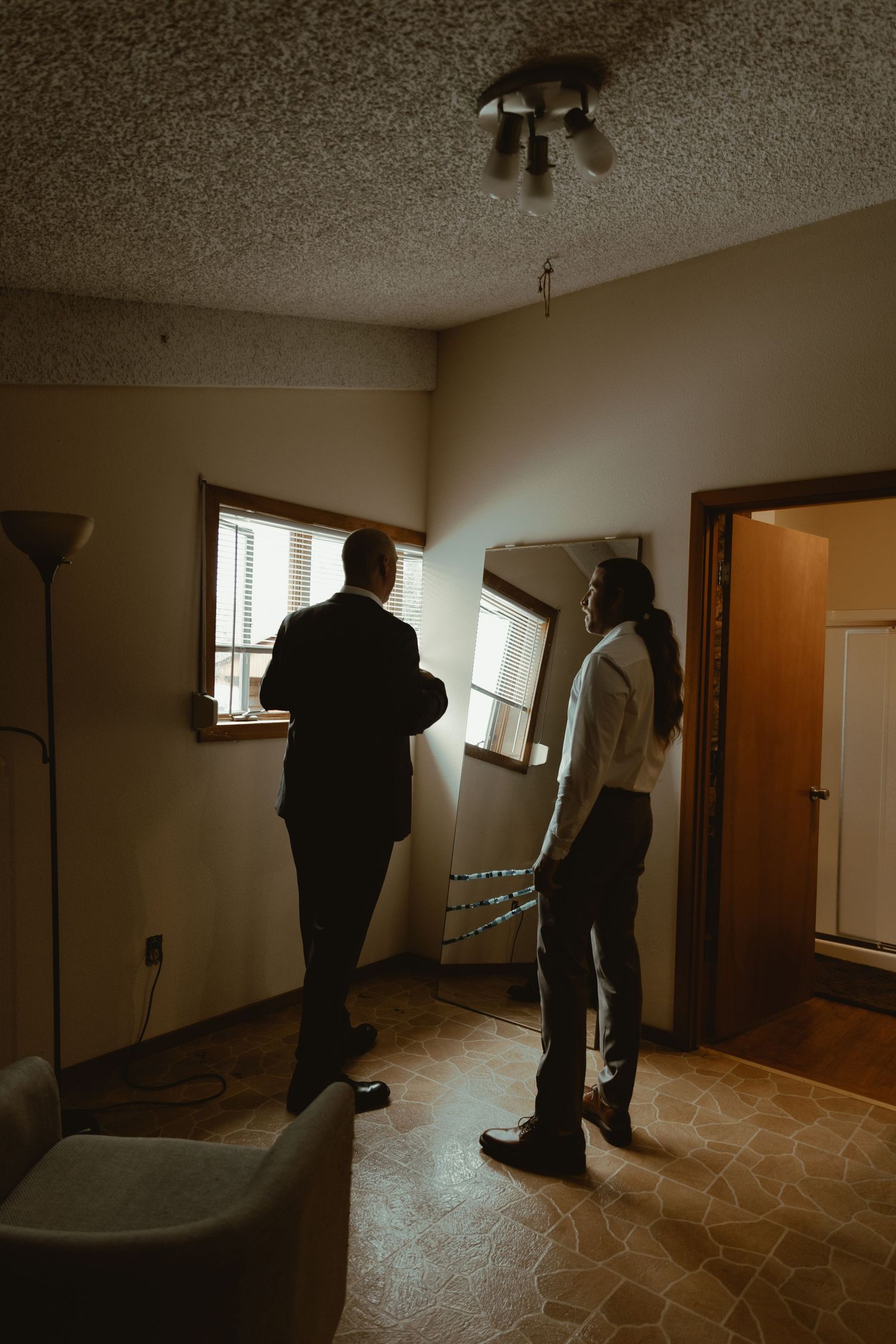 Two formally dressed men stand in silhouette near a window in a softly lit interior room.