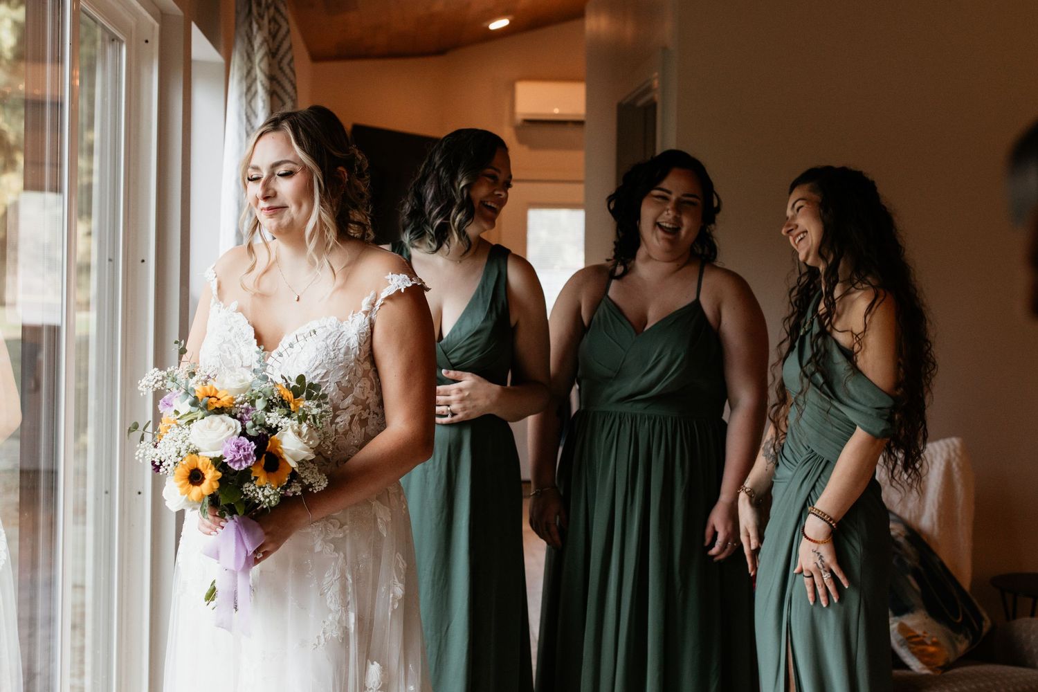 Bride in lace gown holding colorful bouquet laughing with bridesmaids in sage green dresses by a window.
