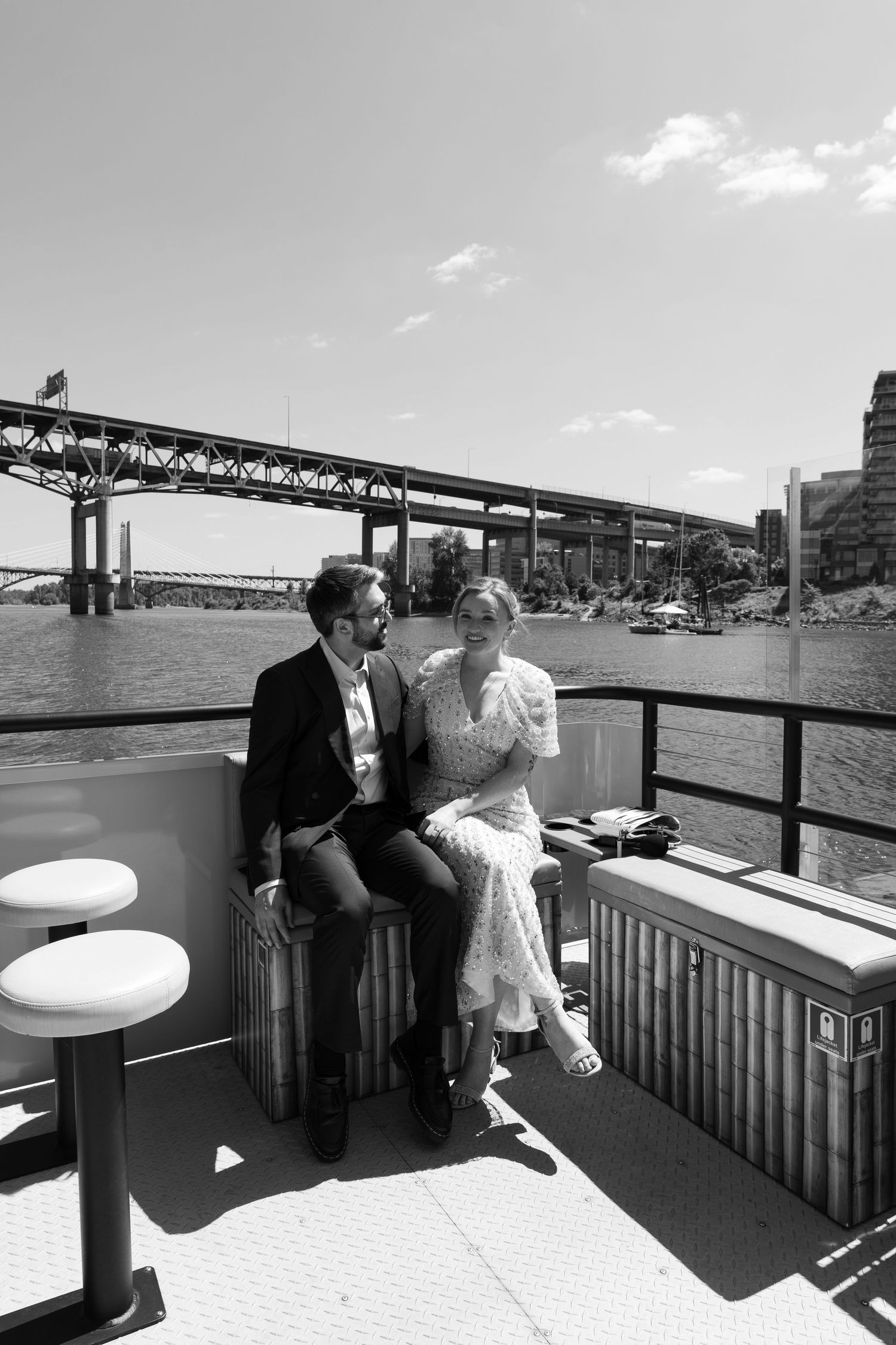 Black and white photo of a couple sitting together on a boat deck with a bridge and river in the background.