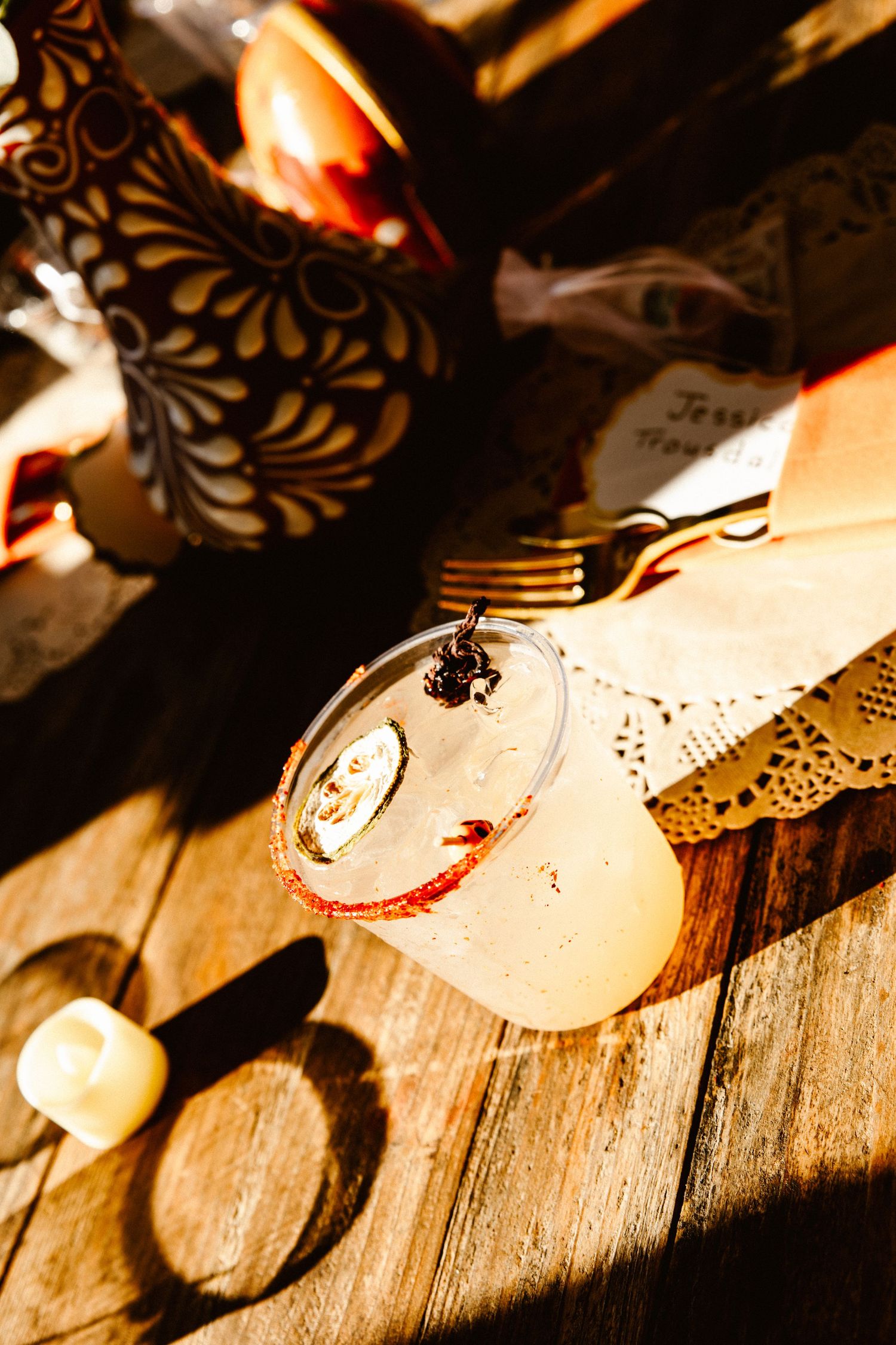 Salt-rimmed cocktail in a clear glass with a garnish, set on a sunlit rustic wooden table beside a painted pitcher, fork and lace doily.